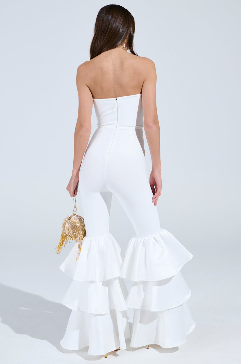 A woman wears the "I Just Wanna Dance Flare Leg Jumpsuit in White" featuring a strapless neckline, layered ruffles, and bell-bottom flares. Facing away, she holds a gold circular clutch with fringes as her long hair cascades down her back against a plain white background.