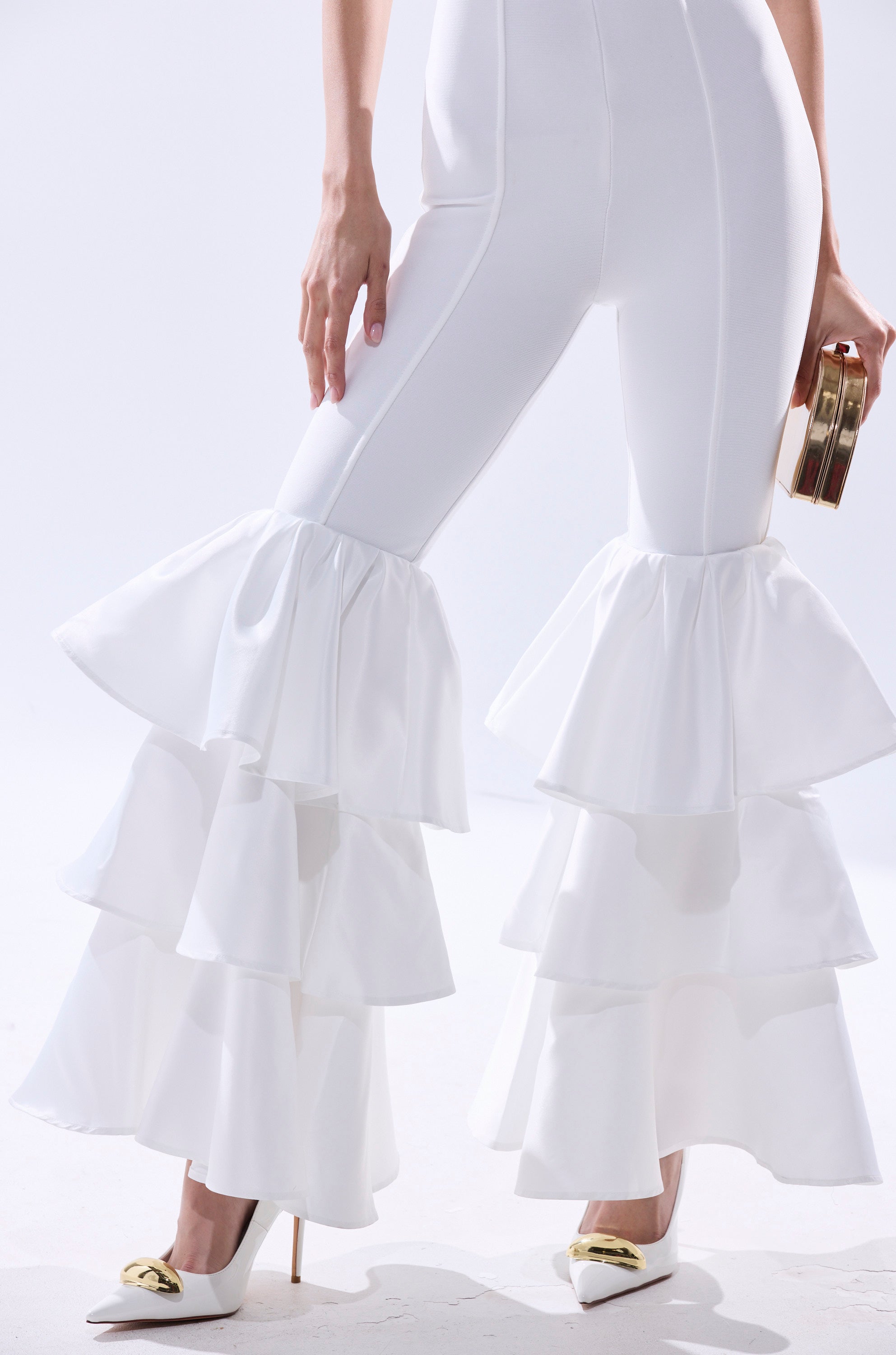 A person stands against a plain white background wearing the I JUST WANNA DANCE FLARE LEG JUMPSUIT IN WHITE with flared legs, white heels with gold accents, and holding a small gold clutch. 4
