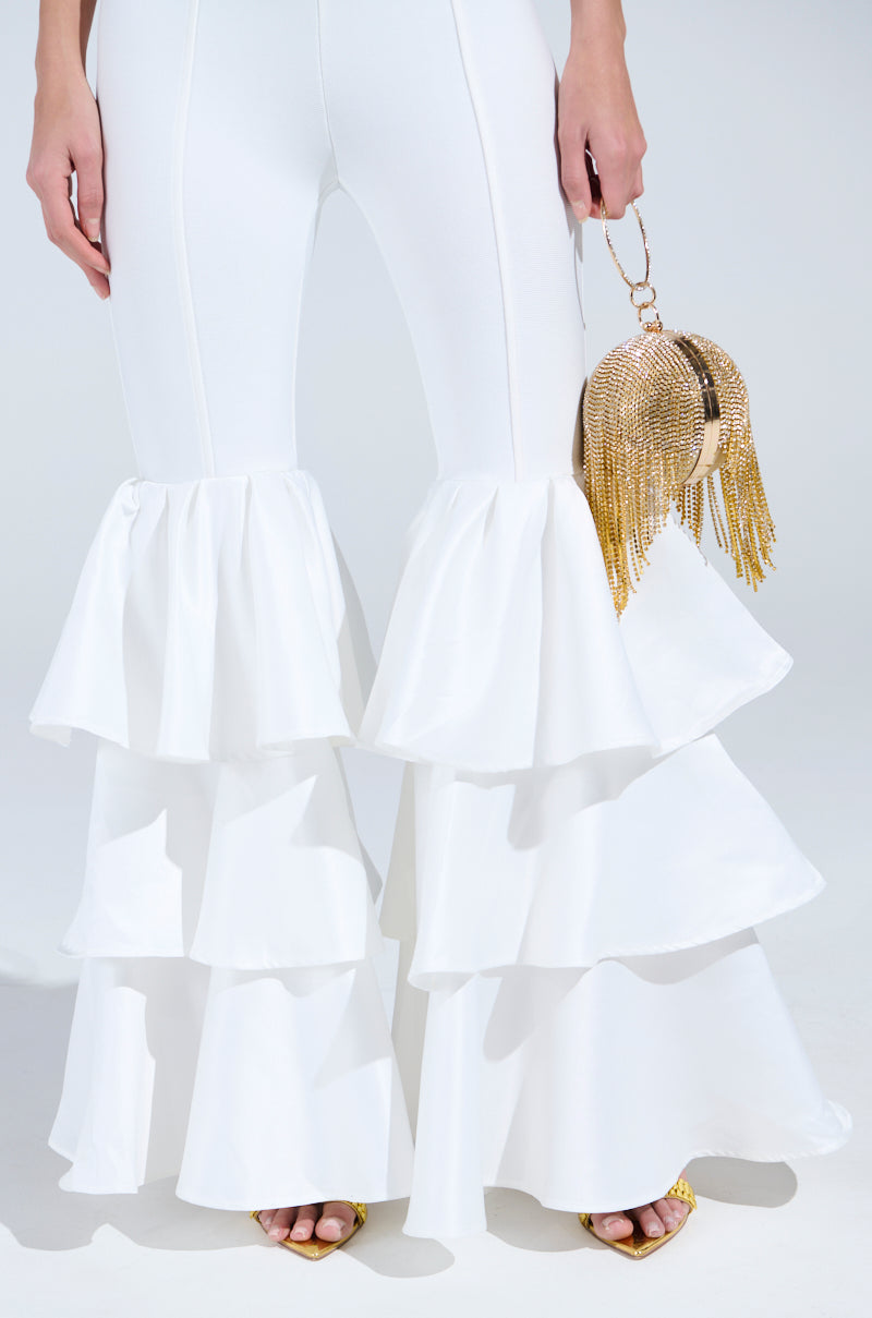 A person wears the I JUST WANNA DANCE FLARE LEG JUMPSUIT IN WHITE, featuring layered ruffles and stretchy fabric, while holding a gold fringed handbag and wearing gold shoes against a plain, light-colored background.