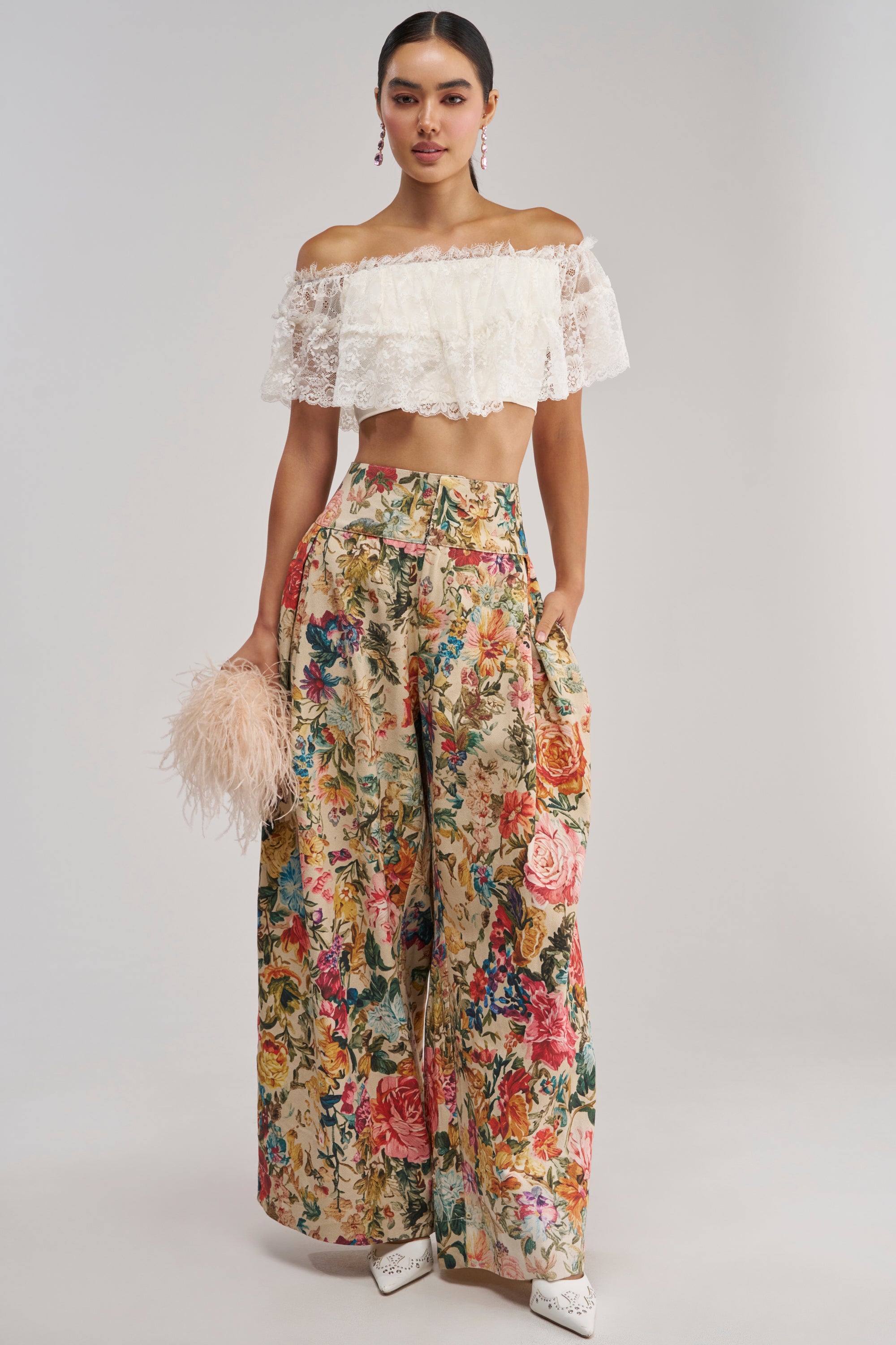A woman stands against a plain background wearing an off-the-shoulder white lace crop top paired with the GIVE ME A FLOWER TROUSER—high-waisted, wide-leg floral print trousers. She accessorizes with a fluffy beige clutch and white shoes.