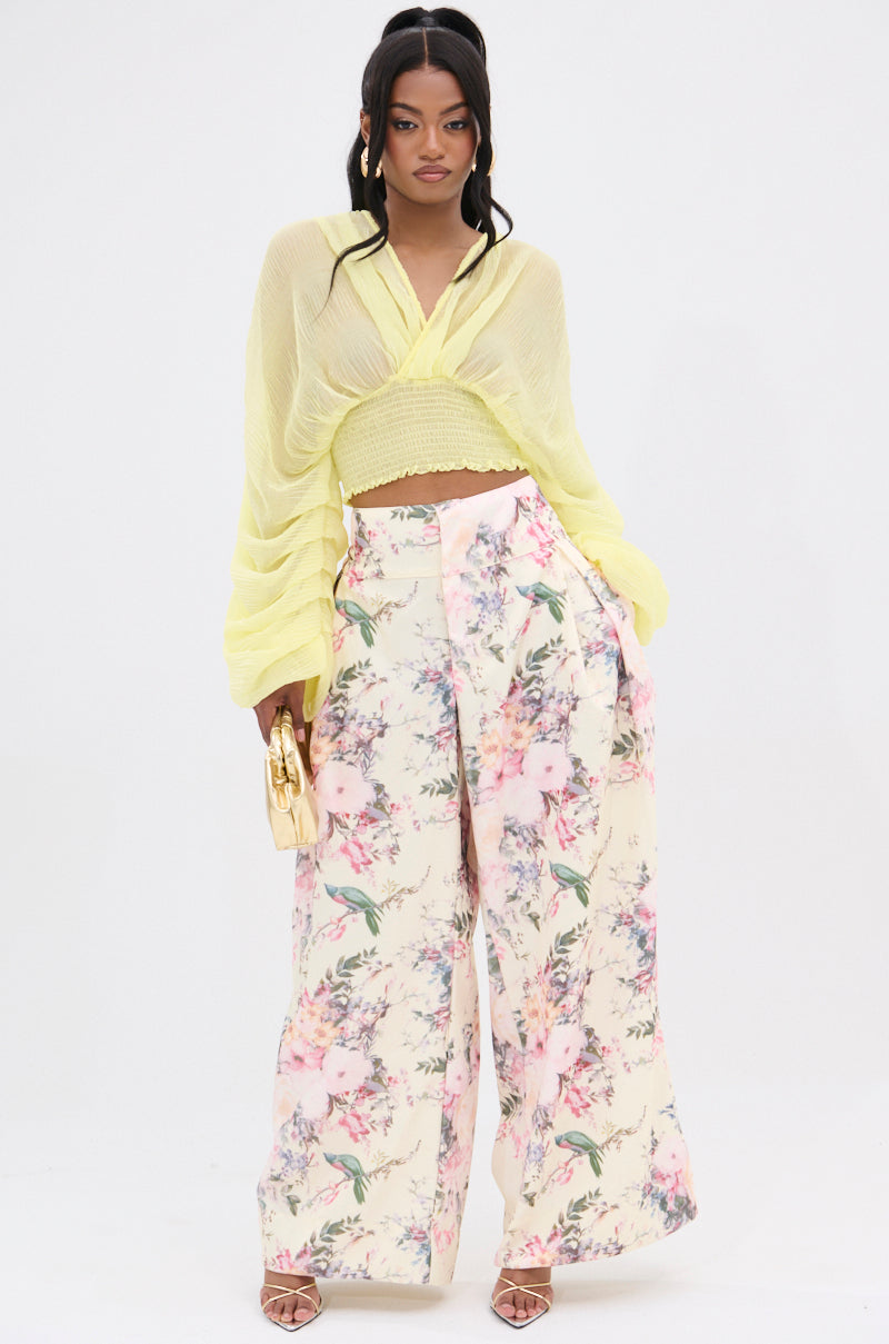 A person models a sheer yellow top over a matching bralette with high-rise SABRINA FLORAL WIDE LEG PANT in a vintage print, complemented by a small gold handbag and strappy heels on a plain white background. 0