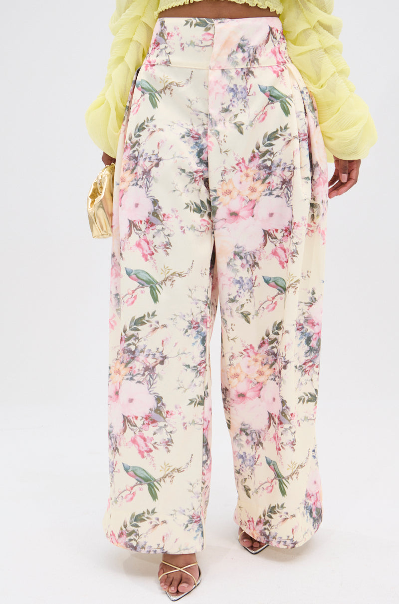 Wearing the SABRINA FLORAL WIDE LEG PANT, a person displays pastel flowers and greenery on a vintage-inspired light background. The high-rise waist pairs well with a textured yellow top as they hold a small gold purse and wear open-toed heels. 1