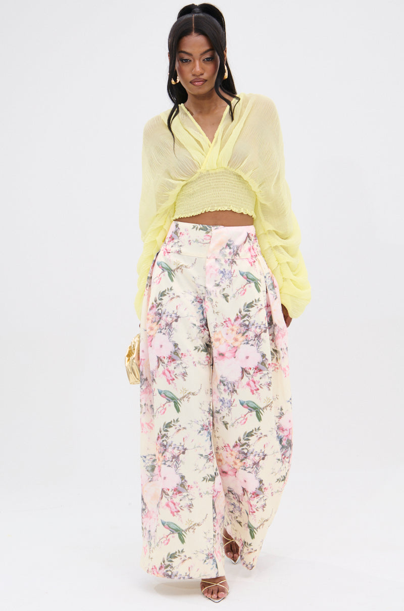 A person wears a sheer yellow blouse with a cropped bandeau top and the SABRINA FLORAL WIDE LEG PANT, showcasing a vintage-inspired print. They have long hair, large hoop earrings, and hold a small handbag against a plain, light-colored background. 6