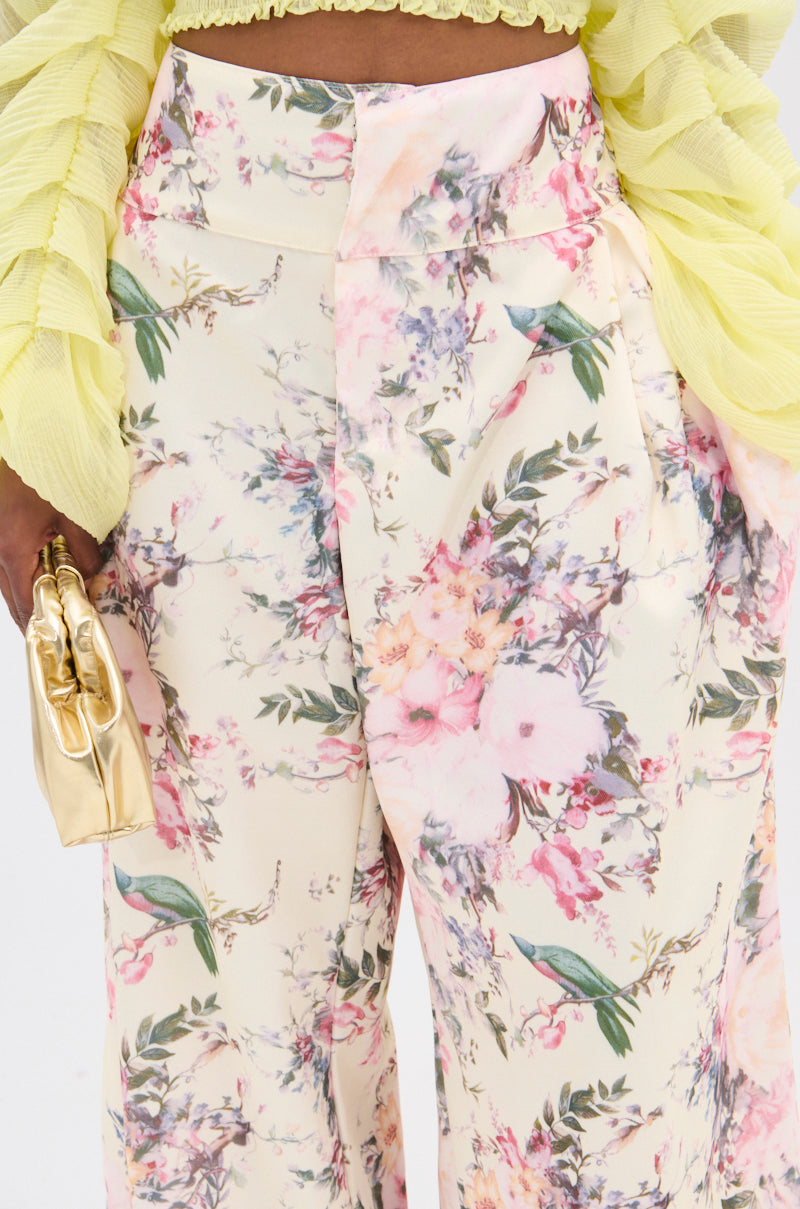 A person dons the vintage-inspired SABRINA FLORAL WIDE LEG PANT with vibrant pink and purple flowers and green leaves. Paired with a light green ruffled top, they hold a small, shiny gold handbag. 7