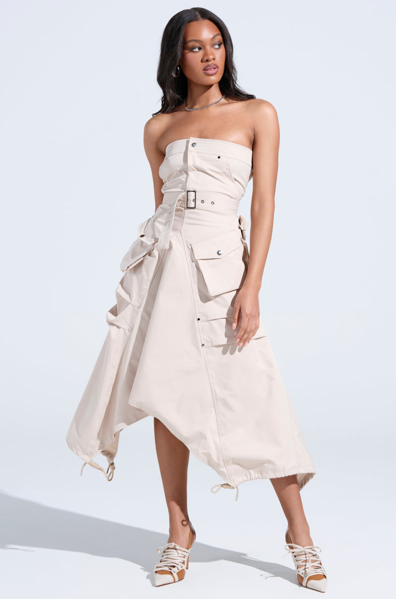 A woman stands confidently in strappy heels against a plain light background, wearing the STROLLIN DOWN SOHO CARGO MIDI DRESS—a strapless design featuring an adjustable waist belt, large pockets, and an asymmetric hem.