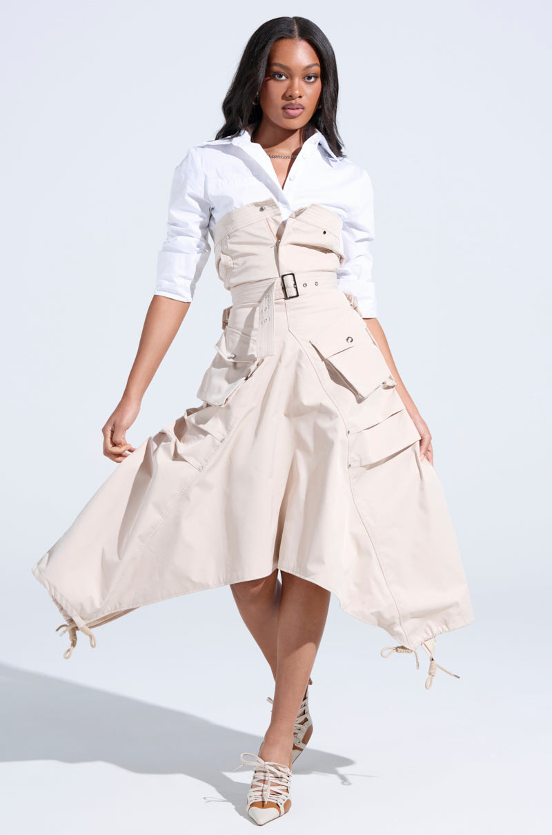 A woman stands against a light background wearing the STROLLIN DOWN SOHO CARGO MIDI DRESS—a beige midi dress with large pockets, adjustable hem, and asymmetrical silhouette—layered over a white shirt and paired with strappy pointed-toe shoes.