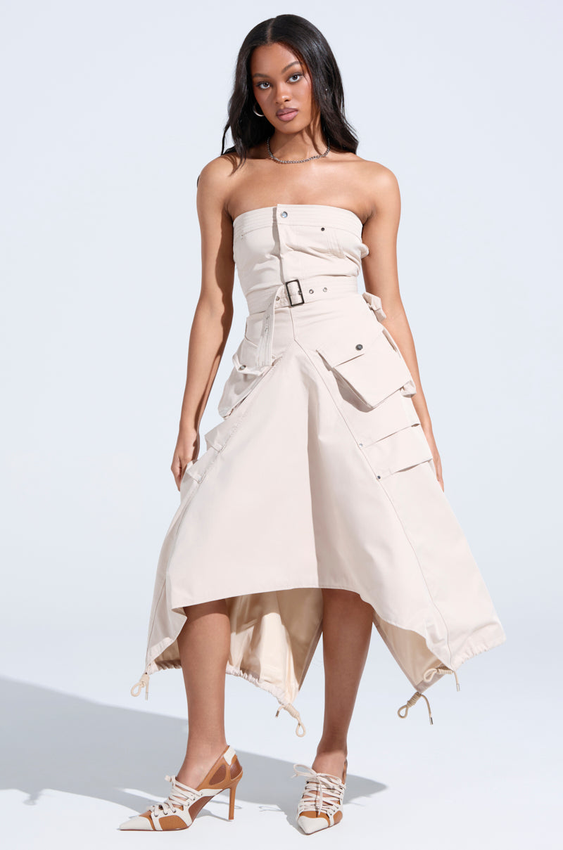 A woman wears the STROLLIN DOWN SOHO CARGO MIDI DRESS—strapless, sculpted bodice, belted waist, large utility pockets, and an asymmetric adjustable hem—paired with pointed high heels against a plain light background.