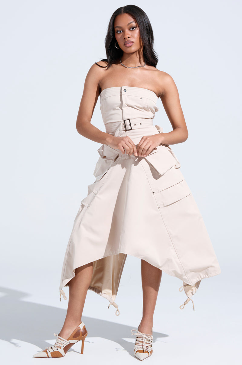 A woman stands confidently in the STROLLIN DOWN SOHO CARGO MIDI DRESS, featuring a sculpted bodice, large pockets, and an asymmetrical hem. She pairs it with strappy pointed-toe heels against a plain light background.