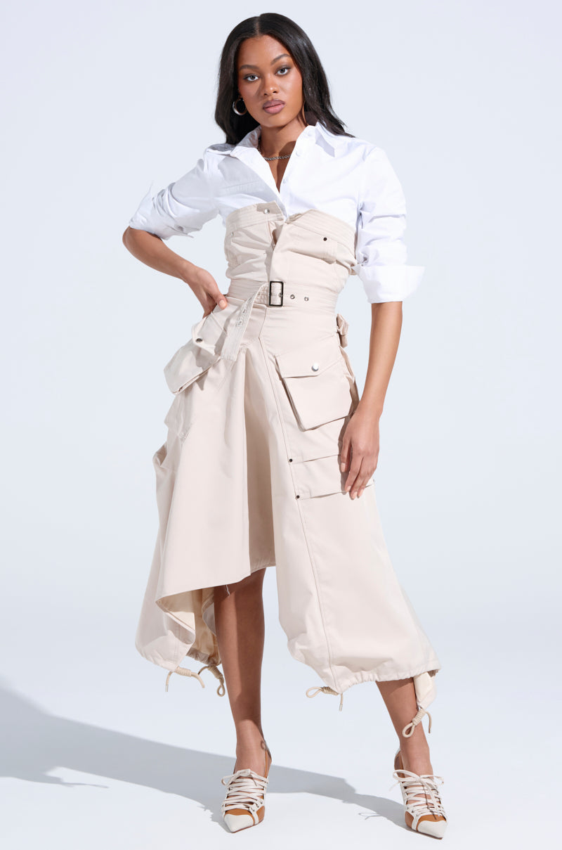 A woman poses in the STROLLIN DOWN SOHO CARGO MIDI DRESS, a white button-up style with beige accents, adjustable hem, sculpted bodice-inspired details, pockets, and a belted waist. She pairs it with strappy heels against a light background.