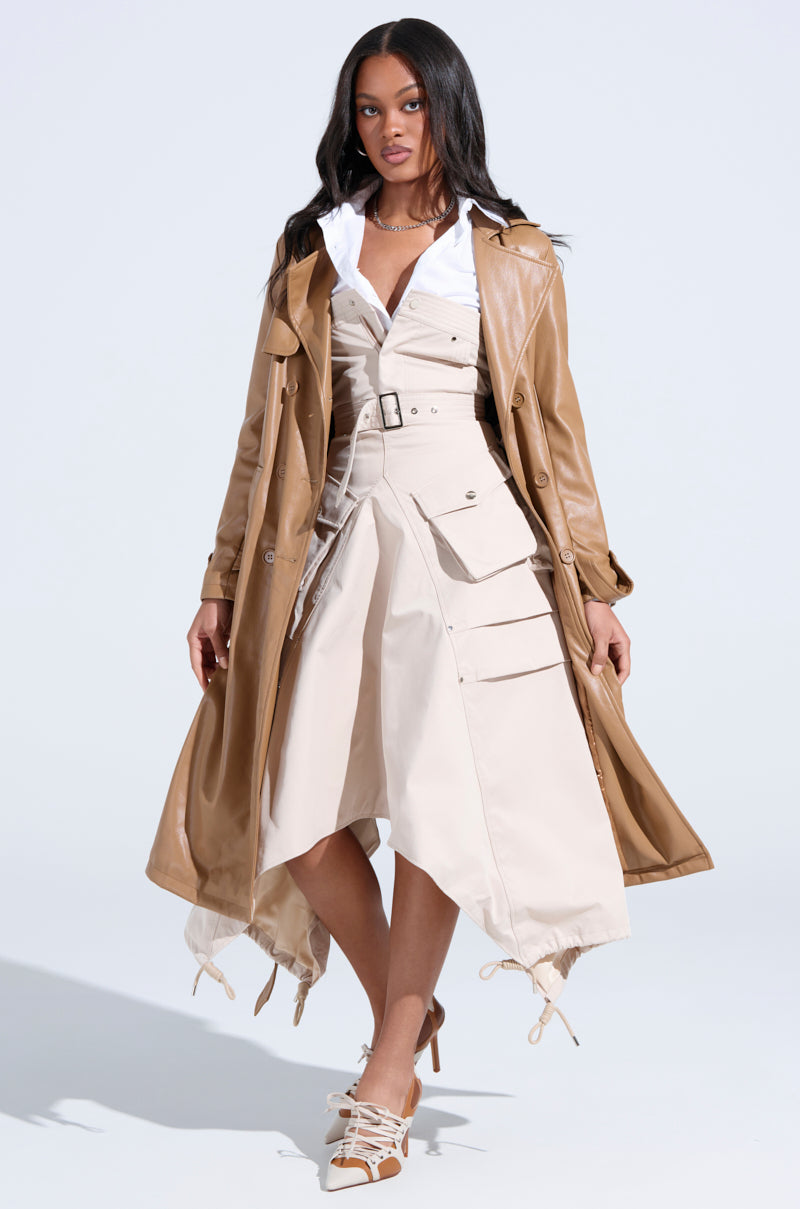 A woman with long dark hair stands against a light background, wearing the STROLLIN DOWN SOHO CARGO MIDI DRESS—a beige midi dress with an asymmetrical adjustable hem and large pockets—layered under a tan leather trench coat and paired with lace-up heels.