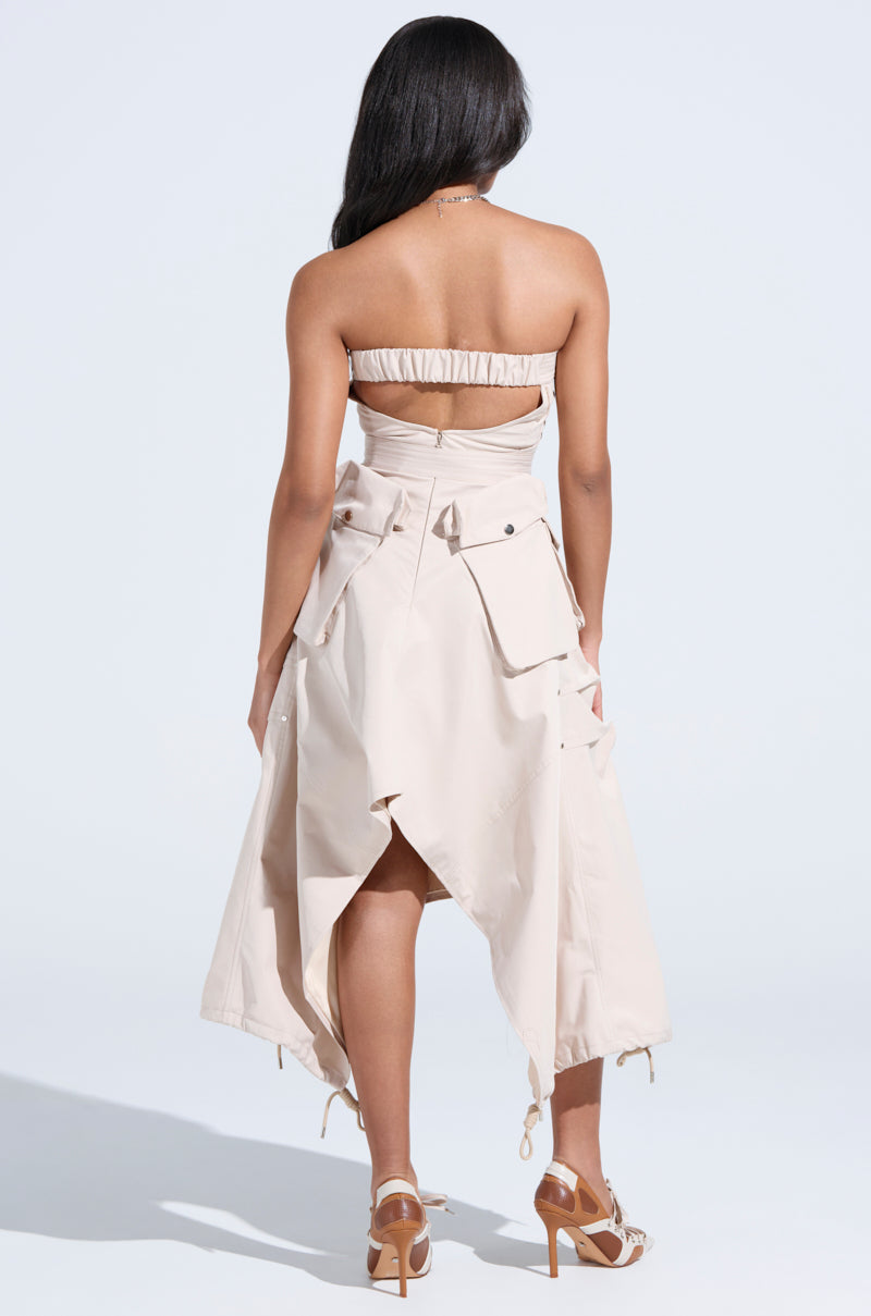A woman with long dark hair faces away, modeling the STROLLIN DOWN SOHO CARGO MIDI DRESS—a beige strapless midi dress with large pockets and an asymmetric hem. She pairs it with high-heeled sandals against a plain, light background.