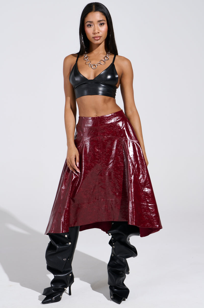 A woman models in a black crop top and the "Think About It" faux leather midi skirt in burgundy. She accessorizes with tall black boots and a chunky chain necklace, all set against a plain white backdrop. 0