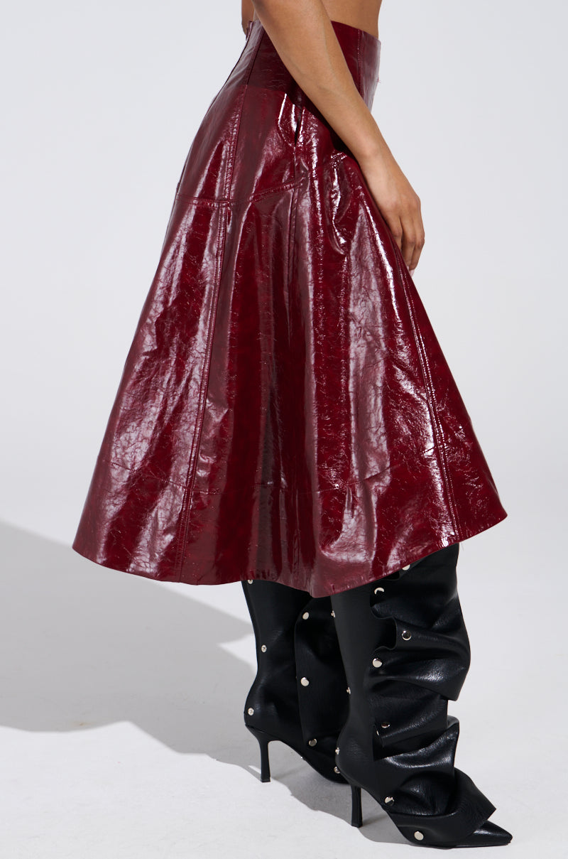 Wearing the THINK ABOUT IT Faux Leather Midi Skirt in Burgundy and black knee-high stiletto boots with decorative studs, a person stands against a plain background. The side view highlights the skirt's rich texture and the boots' intricate design. 3