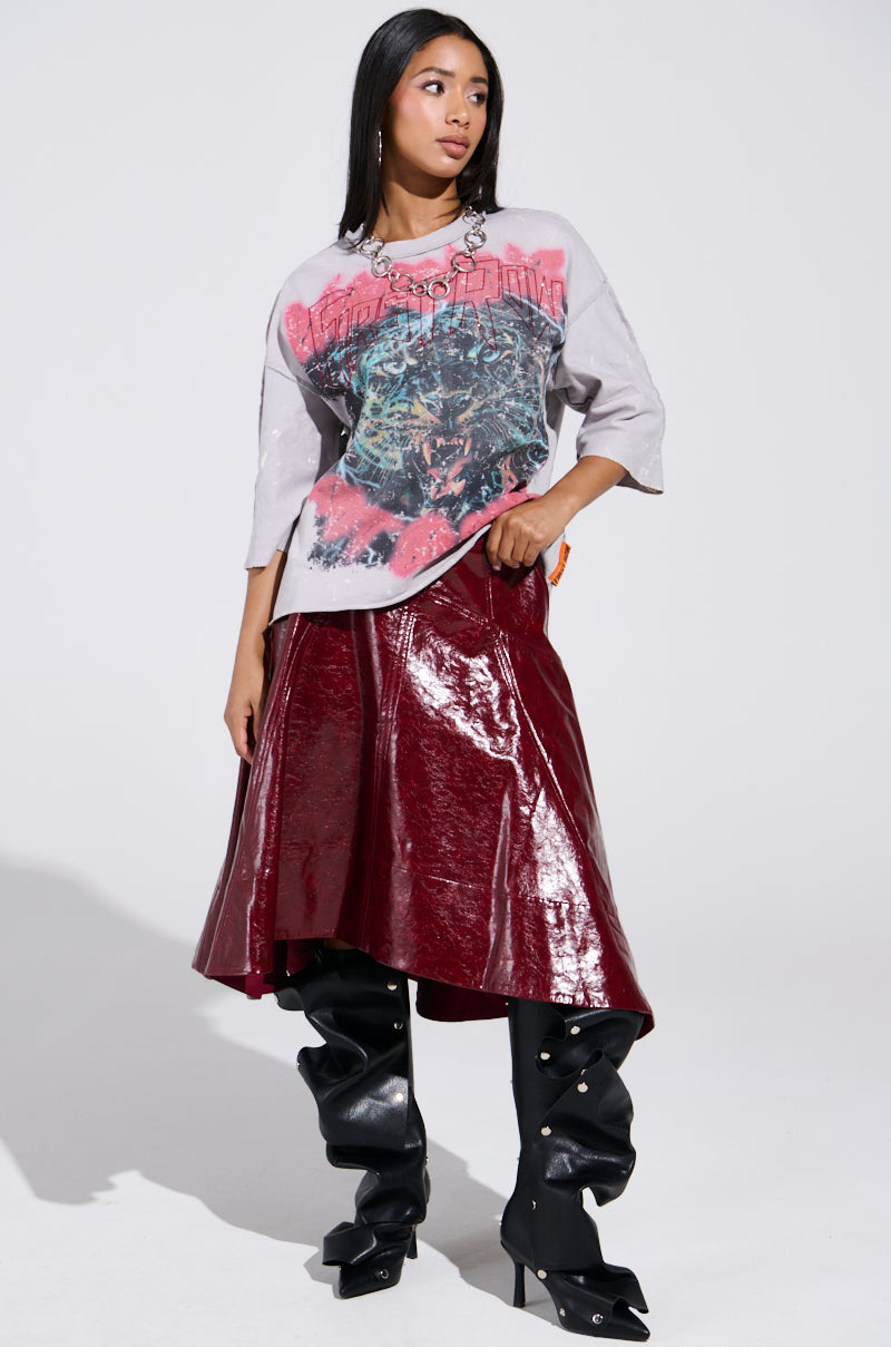 A woman poses in a gray graphic t-shirt and the THINK ABOUT IT FAUX LEATHER MIDI SKIRT IN BURGUNDY, paired with knee-high black boots featuring pearl details. She stands against a white background, looking to the side with one hand on her hip. 6