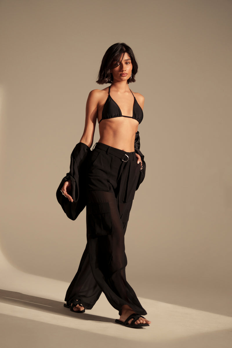 A woman walks confidently in the FAME IS REAL SHEER TROUSER, paired with a black bikini top, wide-leg silhouette, an open black jacket draped over her arms, and black sandals against a neutral studio background.