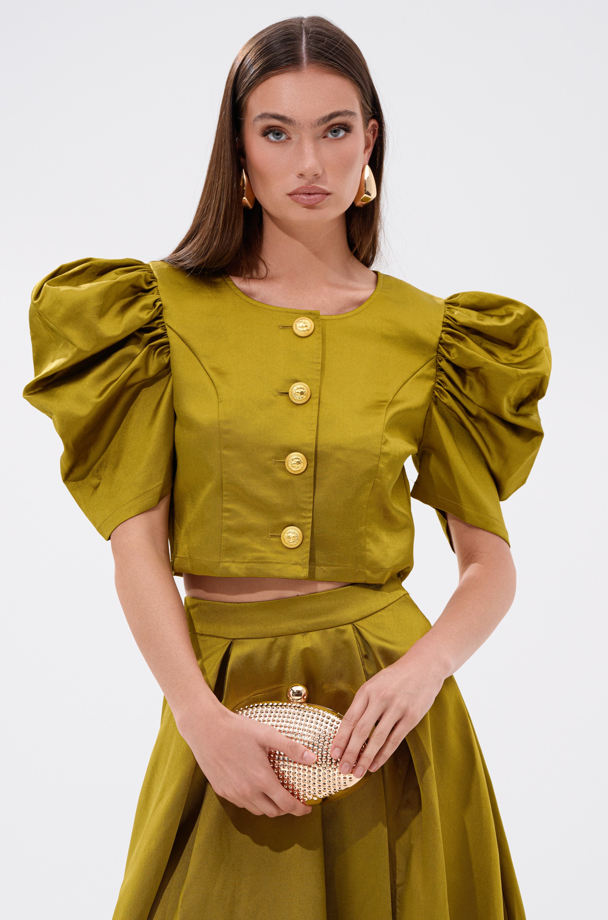 A chic lady wears the CHIC LADY BUTTON DOWN BLOUSE in green satin with gold-tone buttons and puffed short sleeves, paired with a matching skirt. She completes her look with a small gold clutch, long straight hair, and gold drop earrings.
