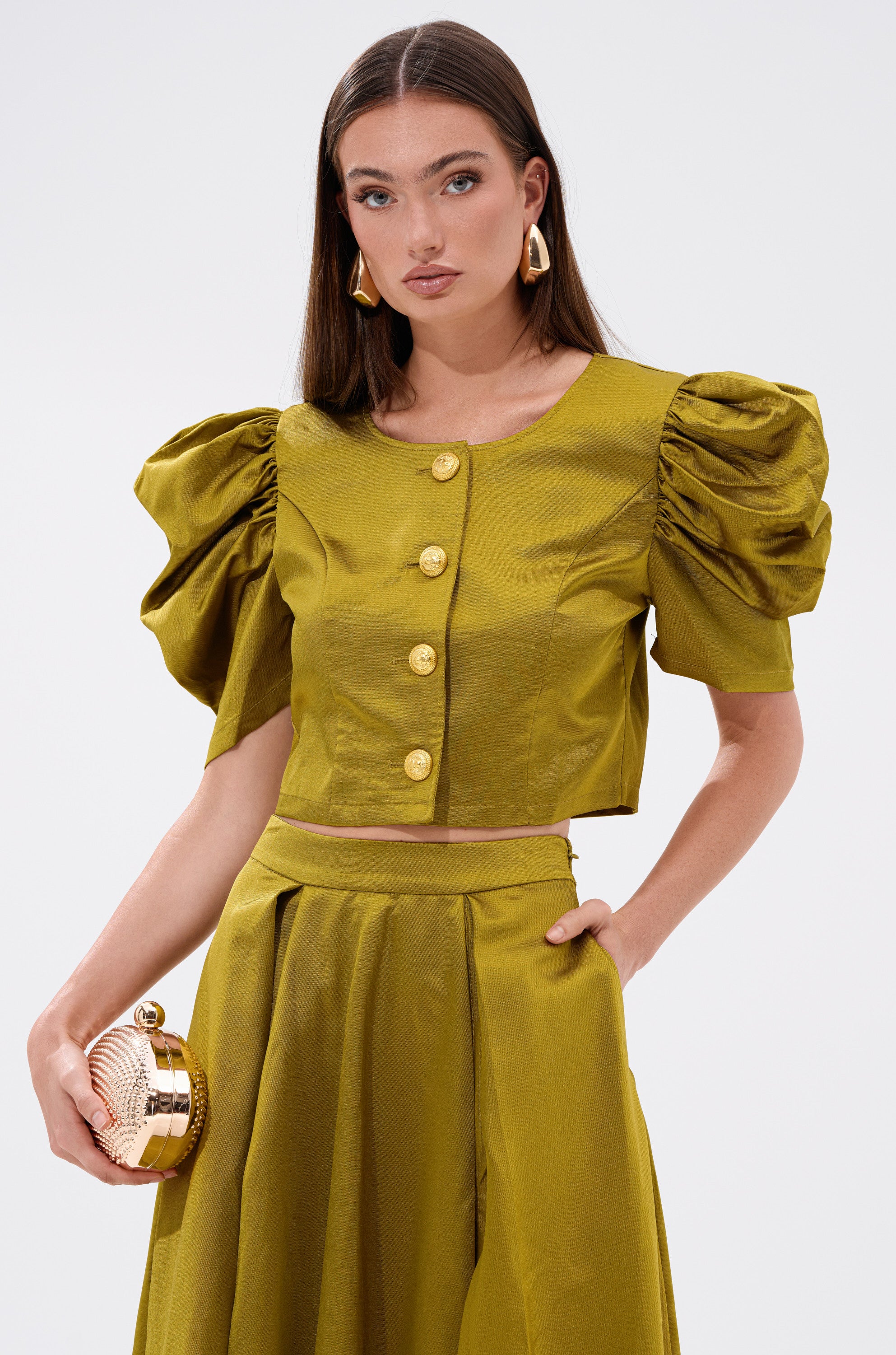 A woman wears the CHIC LADY BUTTON DOWN BLOUSE in shiny olive green, featuring a cropped fit, puffed short sleeves, and large gold buttons. She accessorizes with a small round gold clutch and gold earrings, her long hair styled straight.