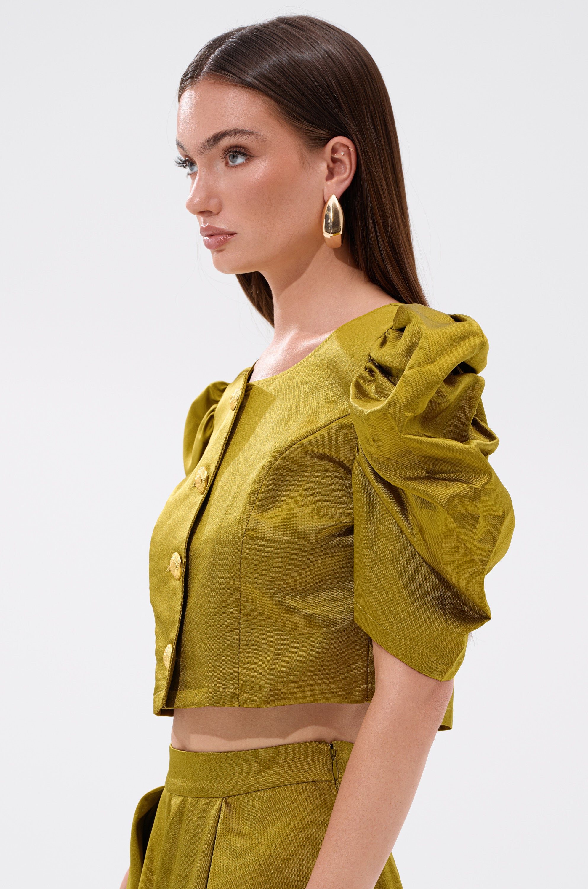 A chic lady models the CHIC LADY BUTTON DOWN BLOUSE, featuring puffed short sleeves, gold buttons, and a cropped cotton-blend silhouette, styled with gold hoop earrings against a plain white background.