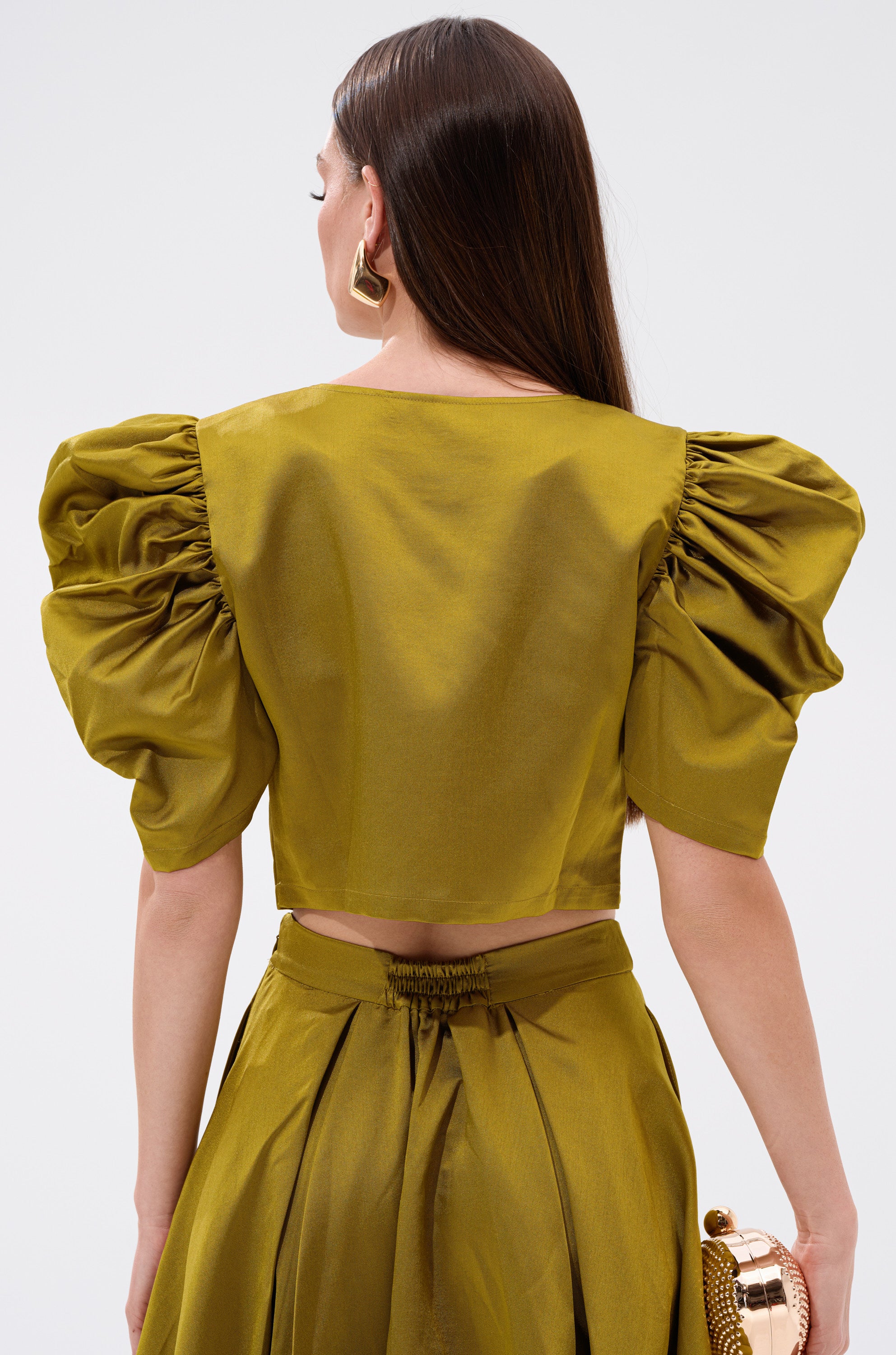 A stylish woman is seen from the back in the CHIC LADY BUTTON DOWN BLOUSE, a glossy olive-green cotton-blend top with puffed short sleeves and a flared skirt. She carries a gold clutch and wears bold geometric earrings for an elevated look.