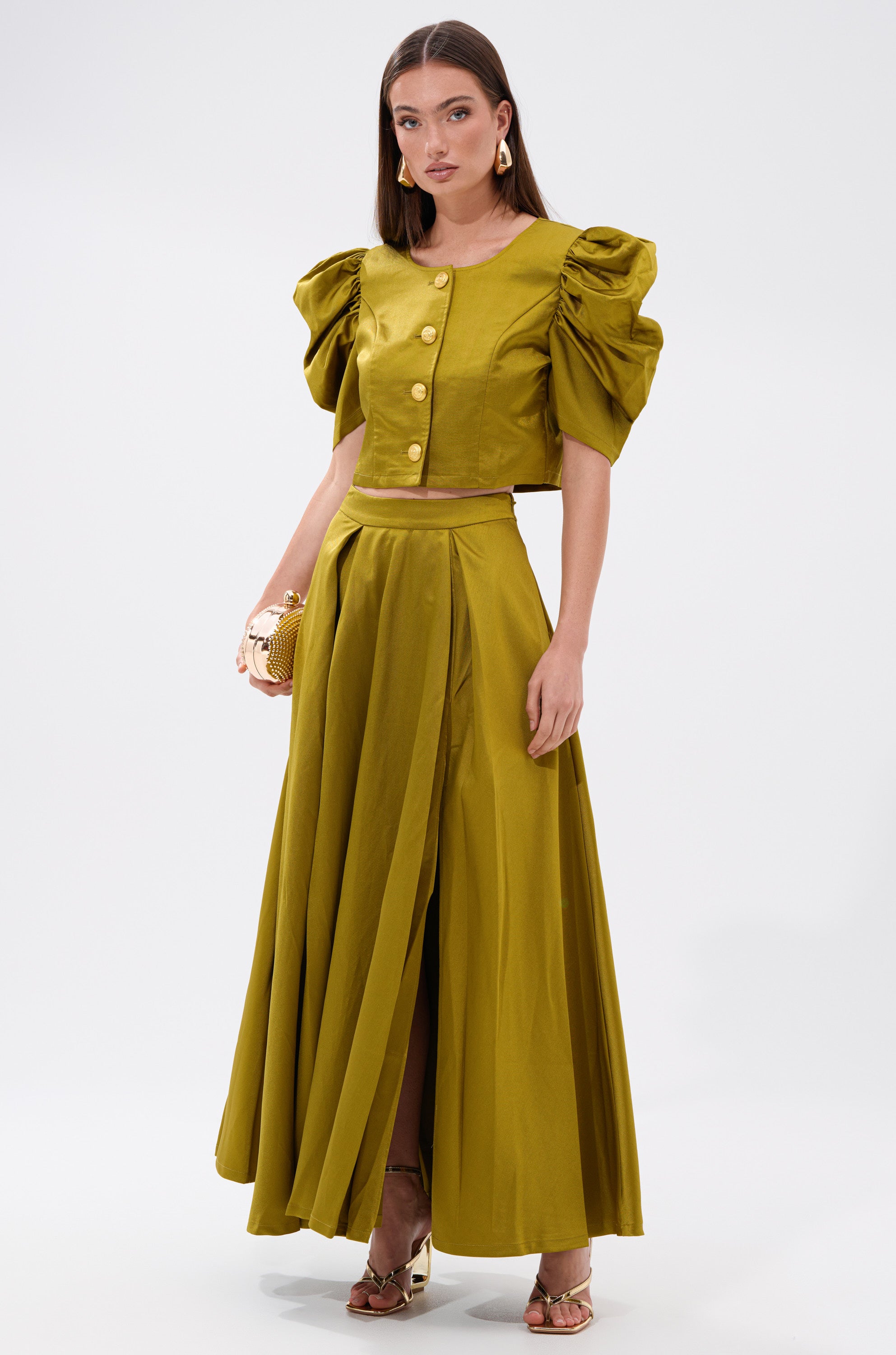 A woman wears the CHIC LADY BUTTON DOWN BLOUSE in olive green, featuring a cropped fit, puffed short sleeves, and gold buttons, paired with a matching front-slit skirt, gold clutch, and gold heels.