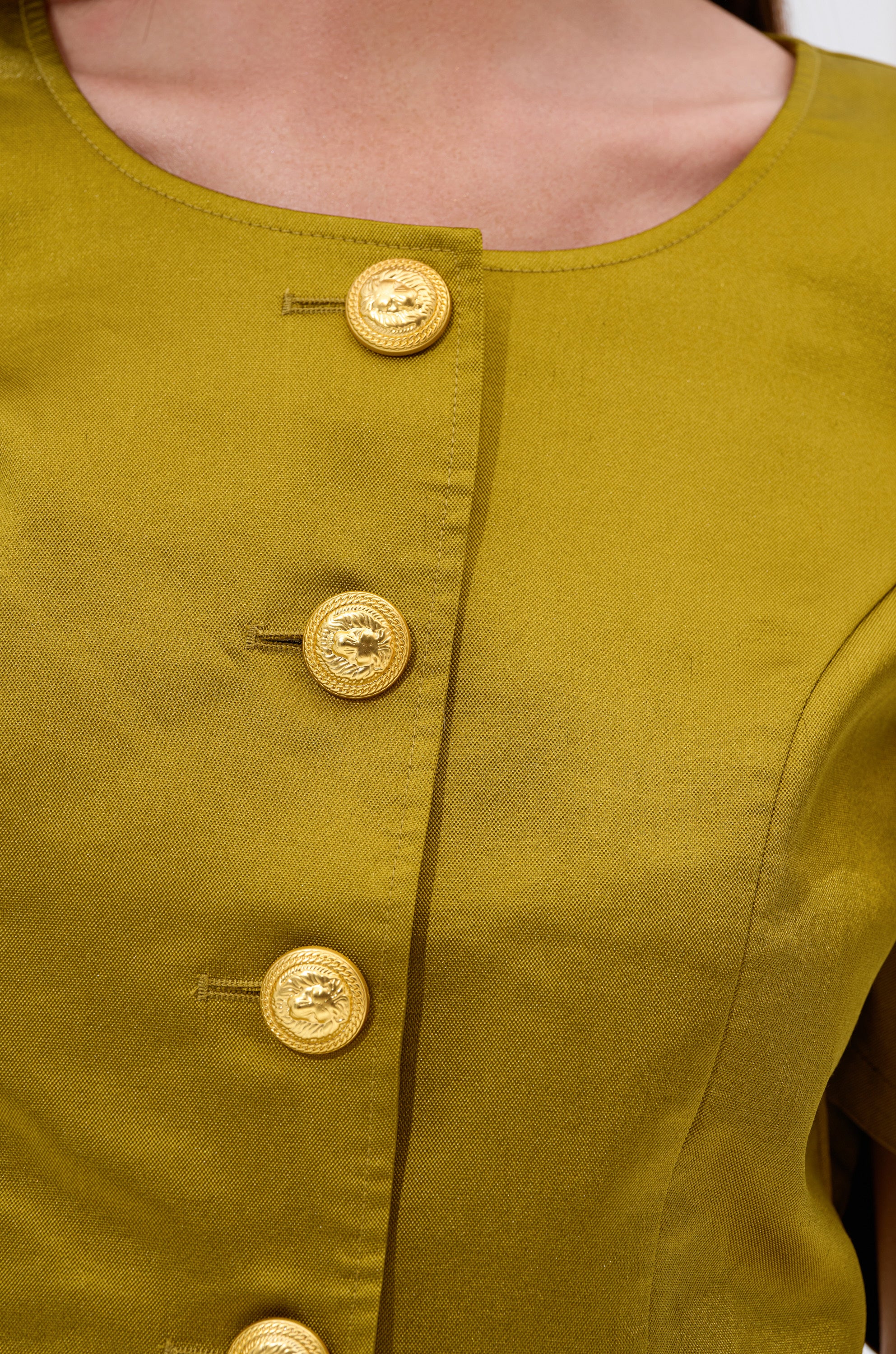 Close-up of a person's upper torso wearing the CHIC LADY BUTTON DOWN BLOUSE in mustard yellow, featuring four gold decorative buttons, a round neckline, and a smooth, slightly shiny cotton-blend fabric with a chic cropped silhouette.
