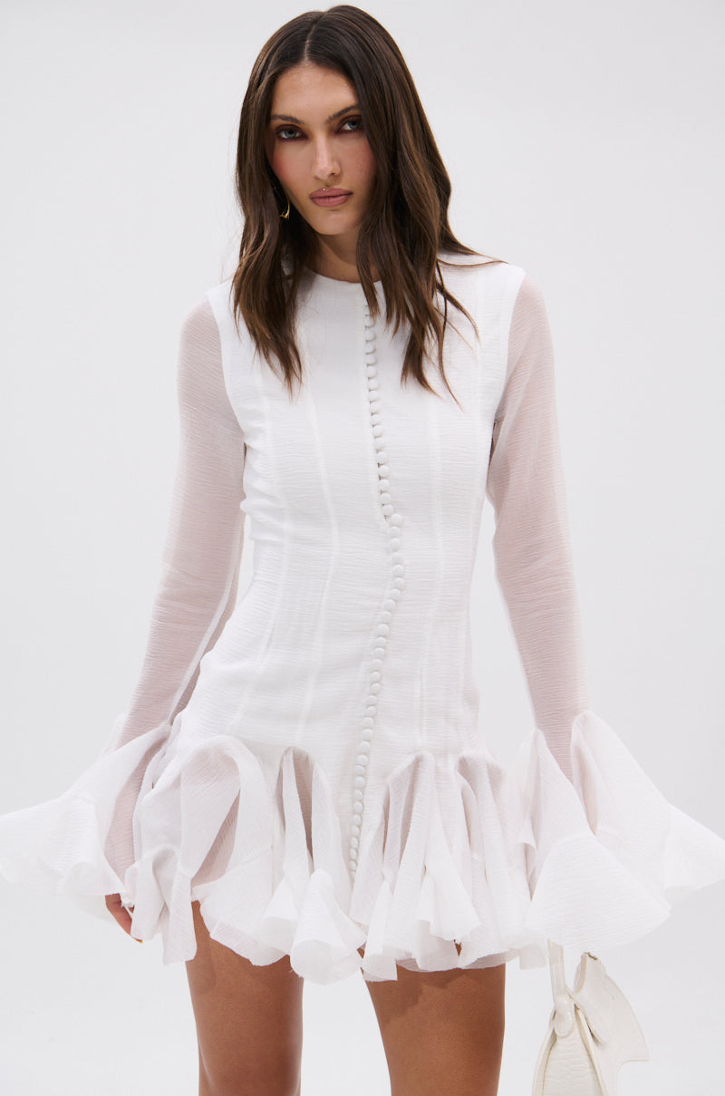 A woman stands against a plain light background wearing the HOURS IN SILENCE RUFFLED MINI DRESS—lightweight and semi-sheer with long sheer sleeves and a ruffled hem. She holds a white handbag, her long dark hair loose, expression neutral.