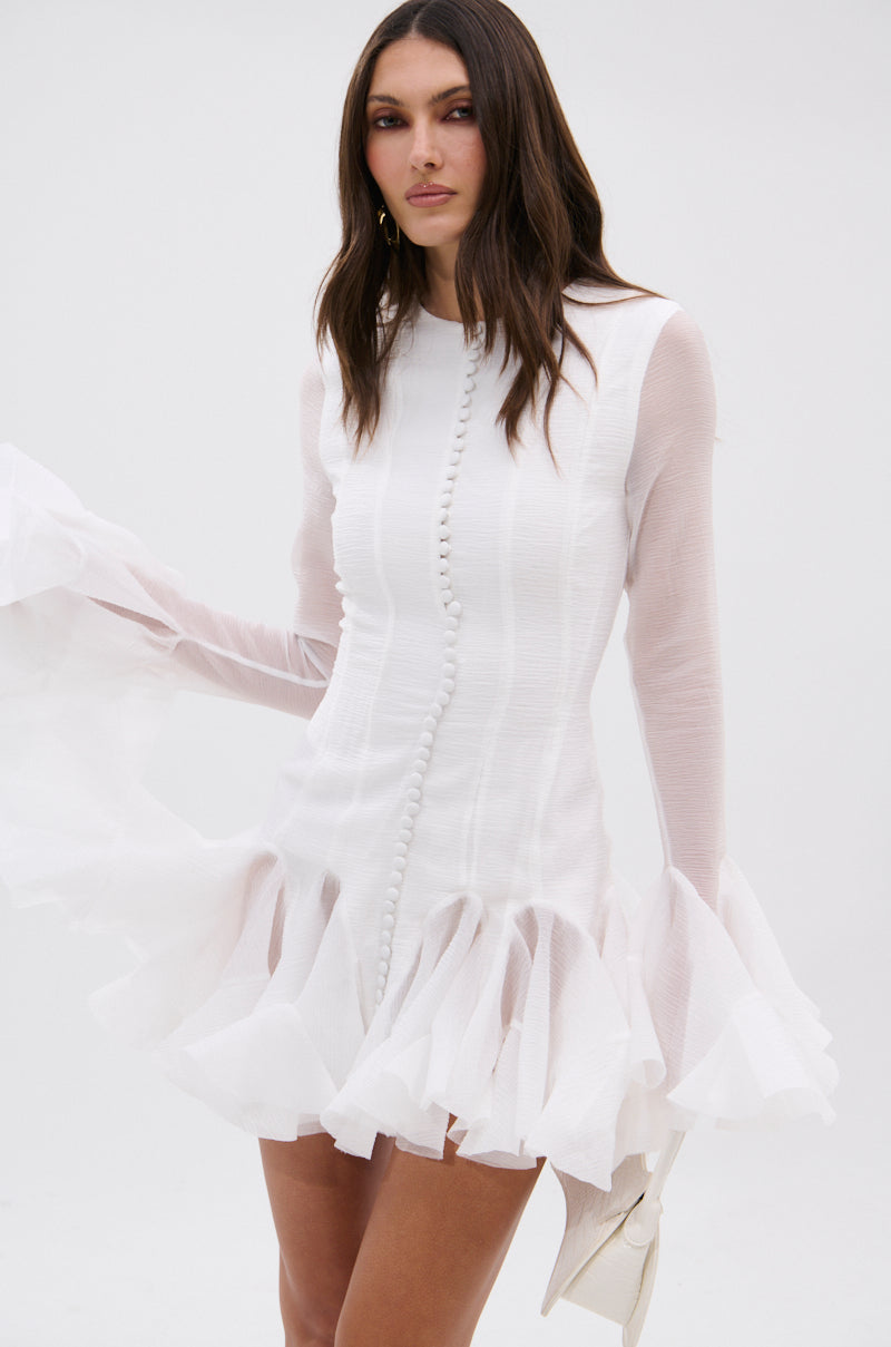 A woman wearing the HOURS IN SILENCE RUFFLED MINI DRESS, a body-skimming white mini with ruffles and a button-down front, stands against a plain backdrop, holding a white clutch and gazing confidently at the camera.
