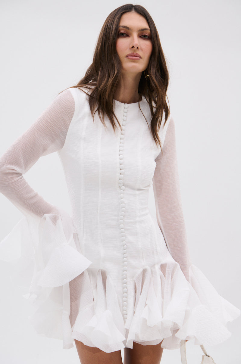 A woman wears the white HOURS IN SILENCE RUFFLED MINI DRESS by AKIRA Label, featuring long sleeves, ruffled cuffs and hem, and fabric-covered buttons. She has long dark hair and stands against a plain light background.