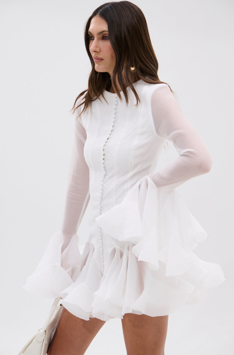A woman stands against a plain light background, holding a light-colored handbag and wearing the AKIRA Label HOURS IN SILENCE RUFFLED MINI DRESS—a white, semi-sheer mini dress with long sleeves, ruffled cuffs and hem, and front buttons.