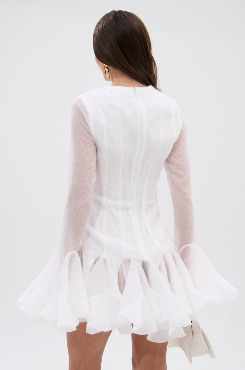 A woman with long brown hair, seen from behind, wears the HOURS IN SILENCE RUFFLED MINI DRESS in semi-sheer textured white fabric and holds a white clutch, standing against a plain light background.