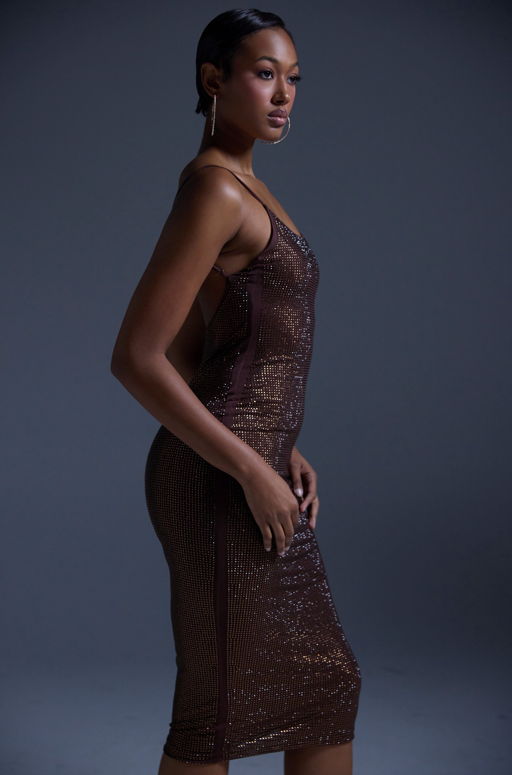 A woman stands in profile against a dark background, wearing the JUST FOR FUN RHINESTONE MIDI DRESS IN BROWN—a curve-hugging, thin-strapped mesh dress. She has short, sleek hair and long, dangling earrings that enhance her glamorous look.