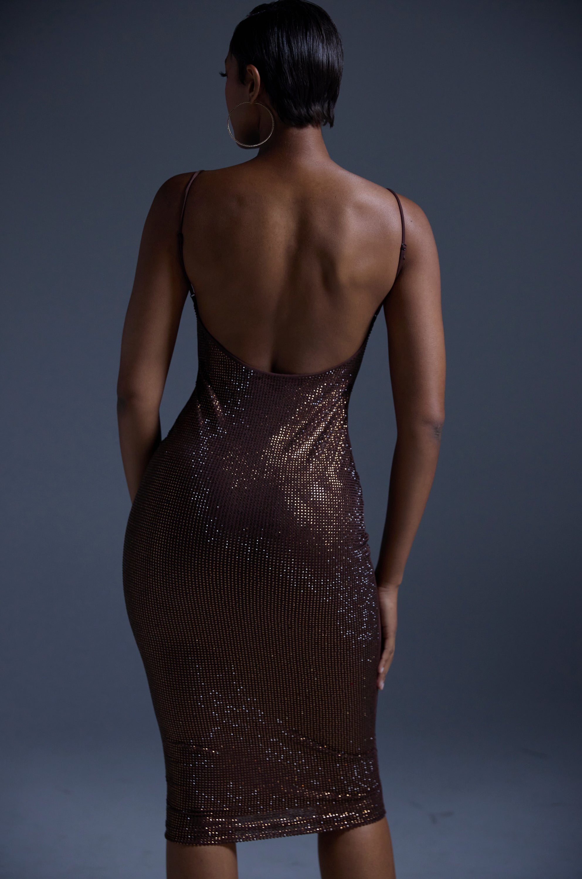 A person models the JUST FOR FUN RHINESTONE MIDI DRESS IN BROWN—a shimmering, curve-hugging mesh dress with thin straps—facing away to reveal their back and short hair against a dark, neutral background.