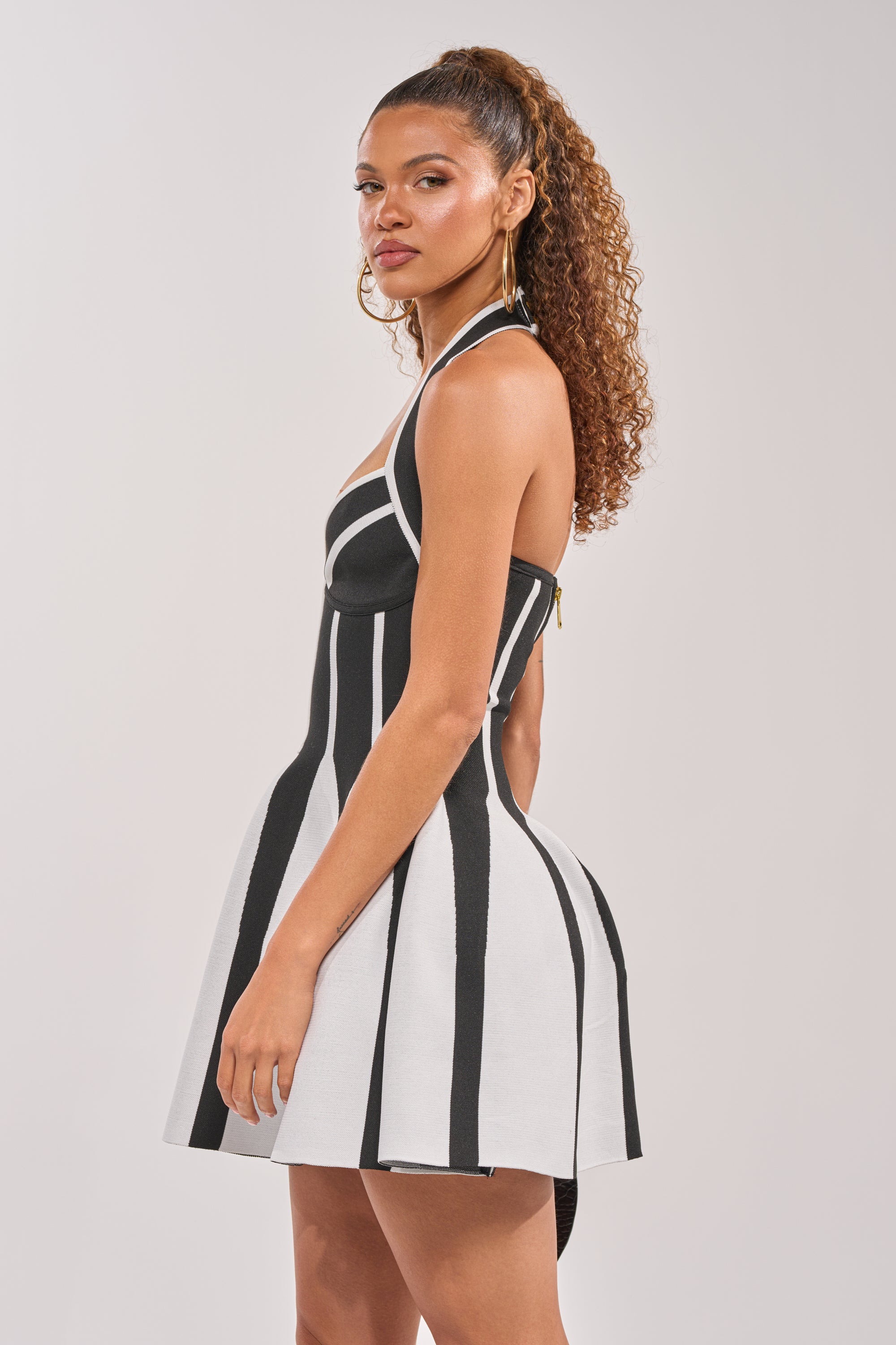 A woman with curly hair in a high ponytail models the PLEASED TO MEET YOU BANDAGE HALTER DRESS, a black and white mini dress with bold vertical stripes, glancing over her shoulder against a plain backdrop. 2