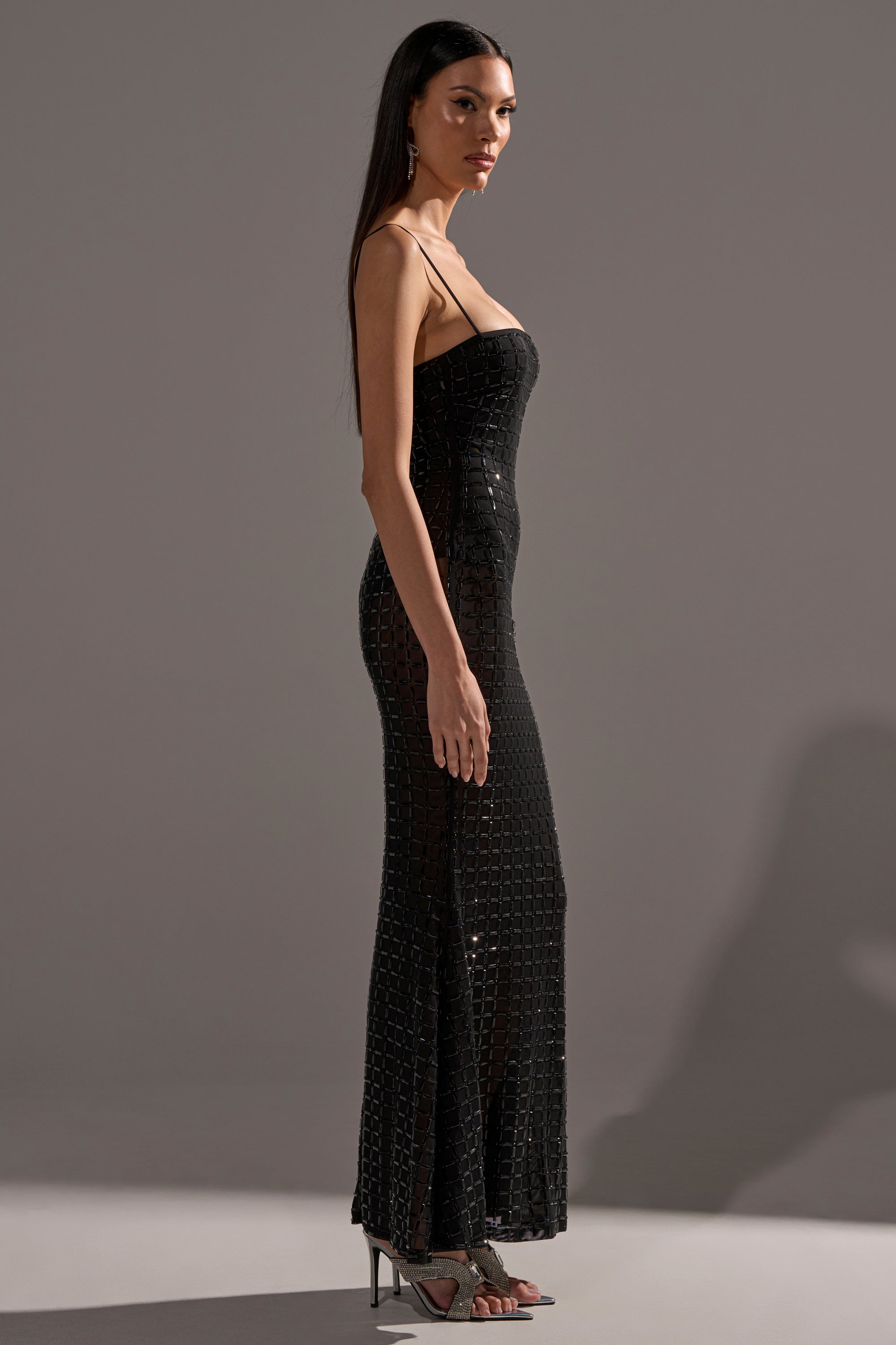 A woman with long dark hair stands in profile, wearing the sleeveless UNTOUCHABLE RHINESTONE MAXI DRESS with thin straps and silver high heels, set against a gray background.