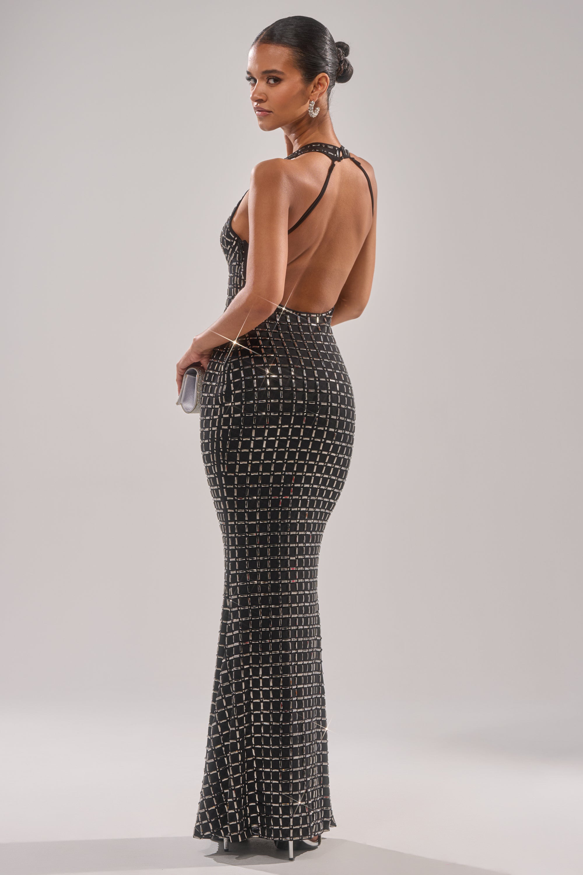 A woman in the DRESSED UP WITH NOWHERE TO GO RHINESTONE MAXI DRESS—floor-length, black with silver geometric rhinestones and a halter neckline—stands hand on hip, looking over her shoulder. She carries a clutch and wears her hair in a sleek bun.