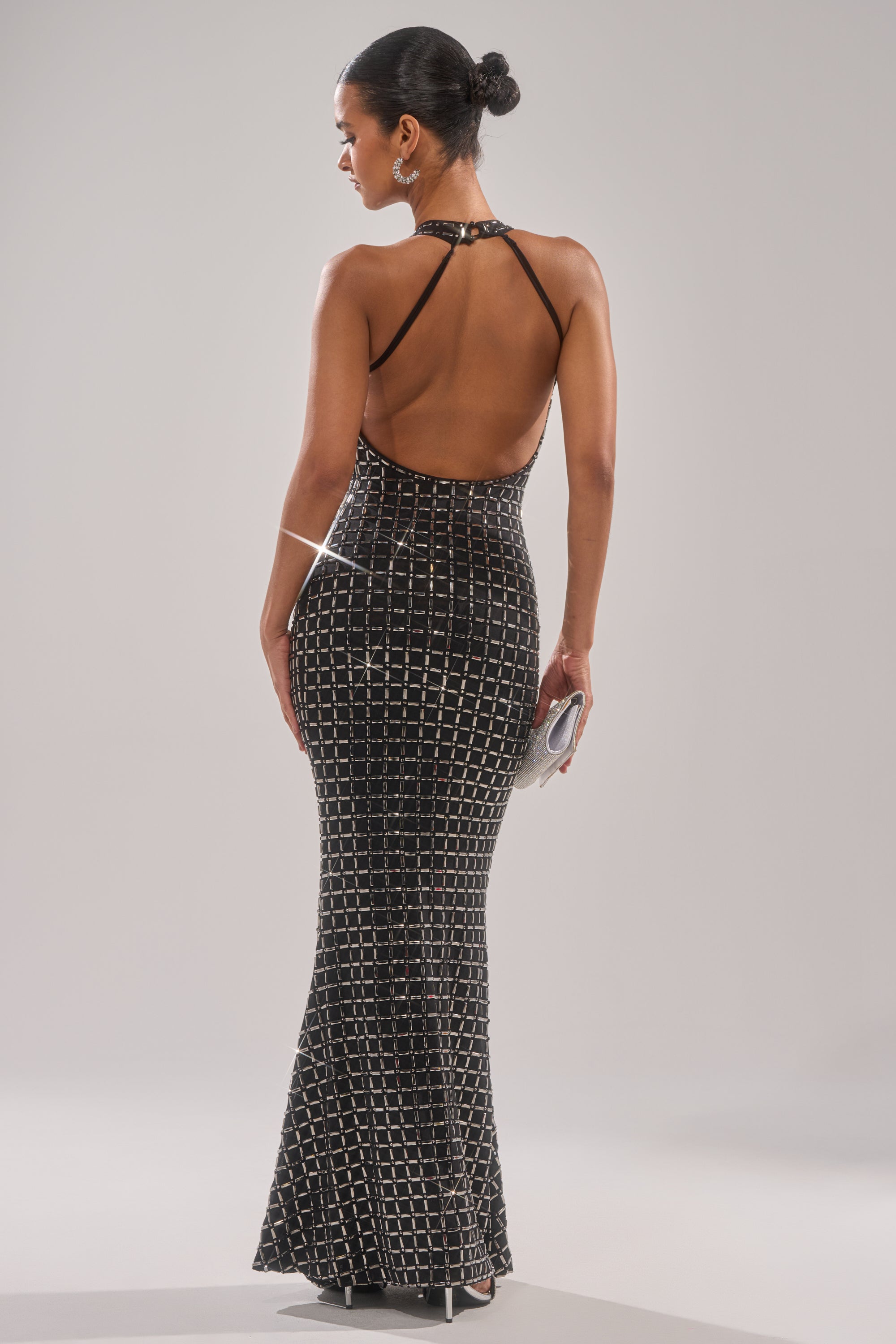 A woman stands facing away in the DRESSED UP WITH NOWHERE TO GO RHINESTONE MAXI DRESS, featuring a sparkling grid pattern, open back, and halter neckline. She holds a clutch and wears her hair in a neat bun.