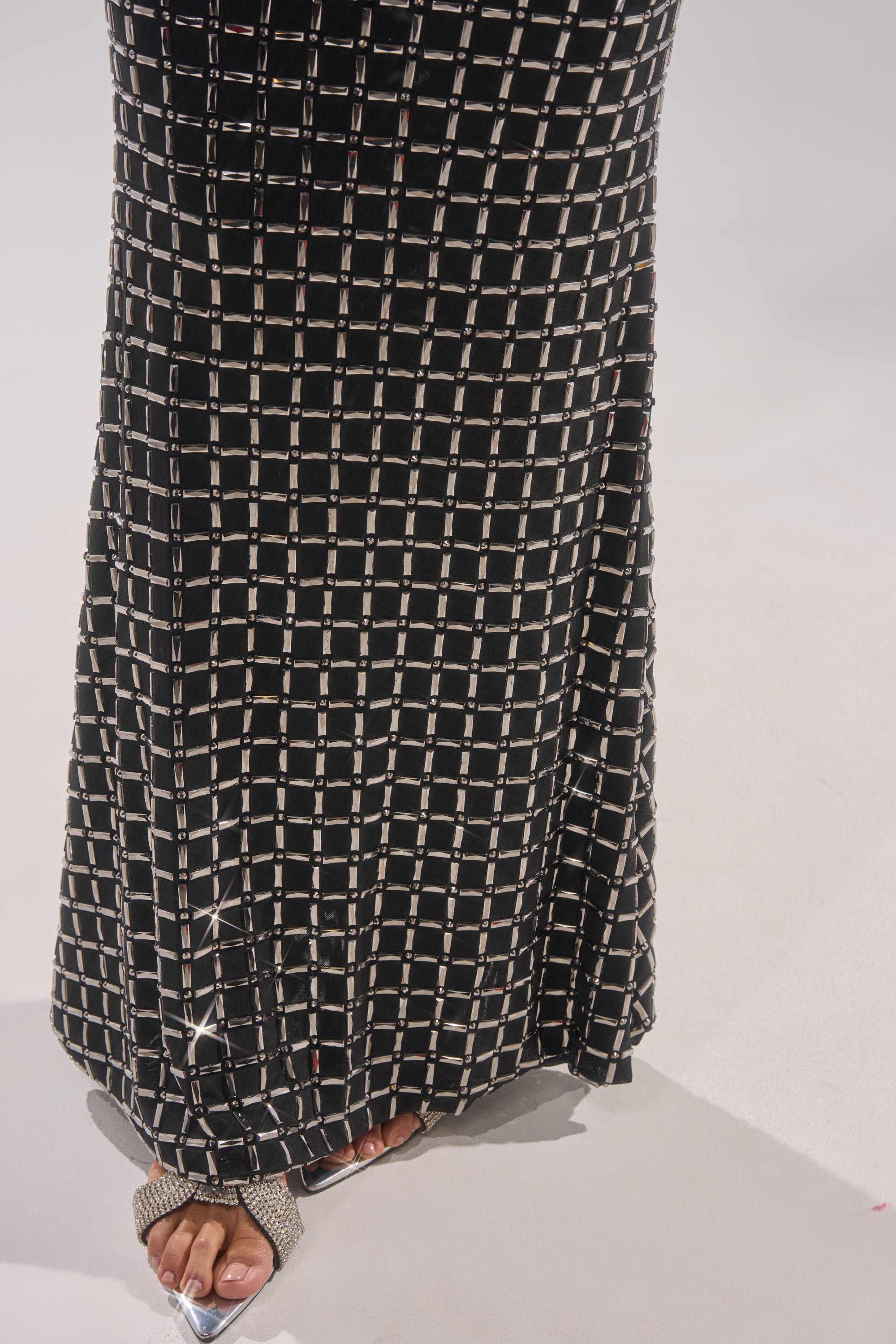 A person stands on a light-colored floor, wearing the DRESSED UP WITH NOWHERE TO GO RHINESTONE MAXI DRESS featuring a black skirt with a white grid pattern and pairing it with open-toe rhinestone sandals. Only lower legs and feet are visible.