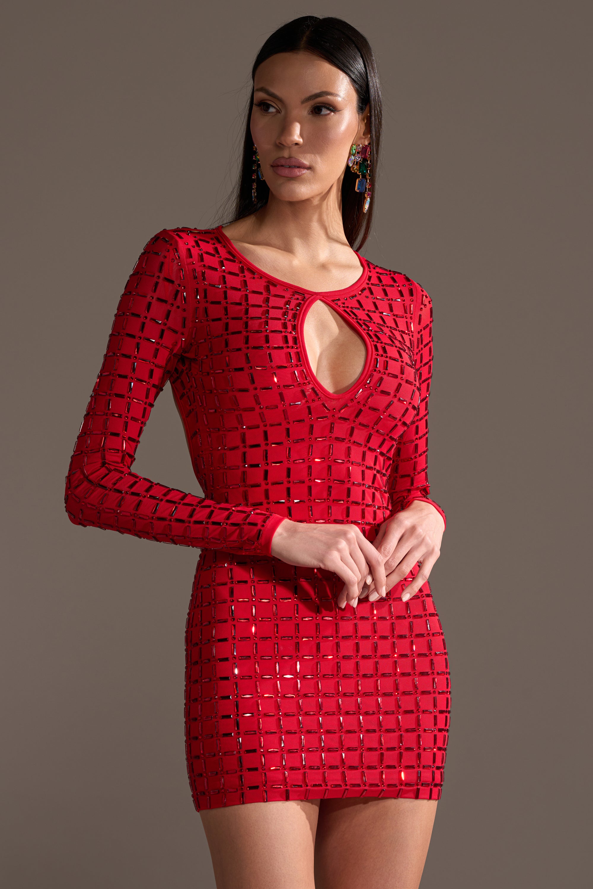 A woman stands against a plain background with her hands clasped, wearing the ALL THAT SHINES RHINESTONE MINI DRESS—a fitted red bodycon featuring long sleeves, a geometric cutout pattern, and a keyhole neckline.