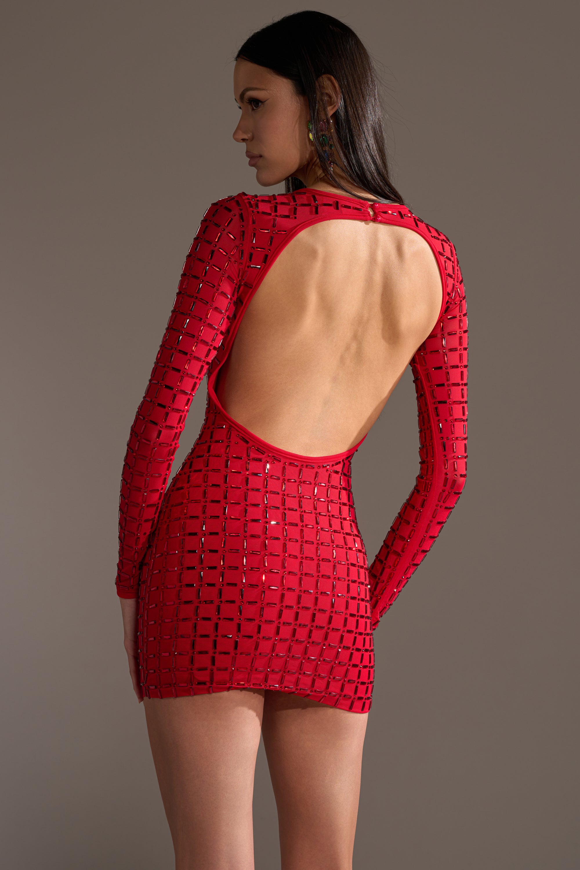 A woman models the ALL THAT SHINES RHINESTONE MINI DRESS—a fitted red bodycon with long sleeves, an open back, and a geometric square pattern—against a neutral background, gazing to the side.