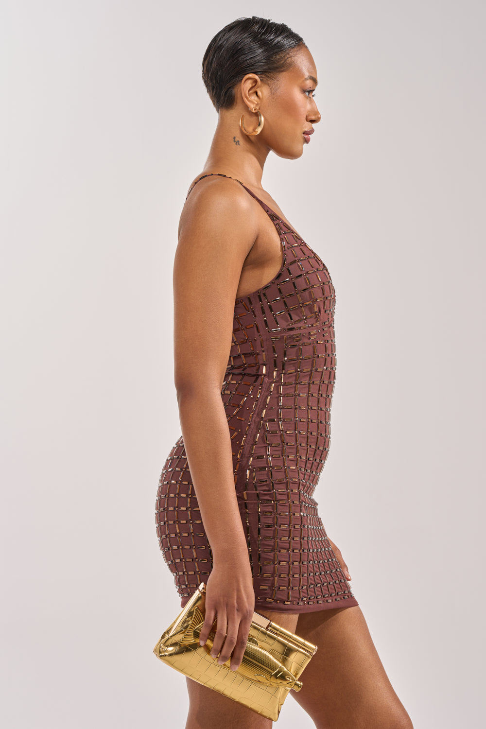 A woman in the DANCE THE NIGHT AWAY RHINESTONE MINI DRESS stands in profile, holding a shiny gold clutch. She has short hair and large hoop earrings, creating a chic, party-ready look. 3
