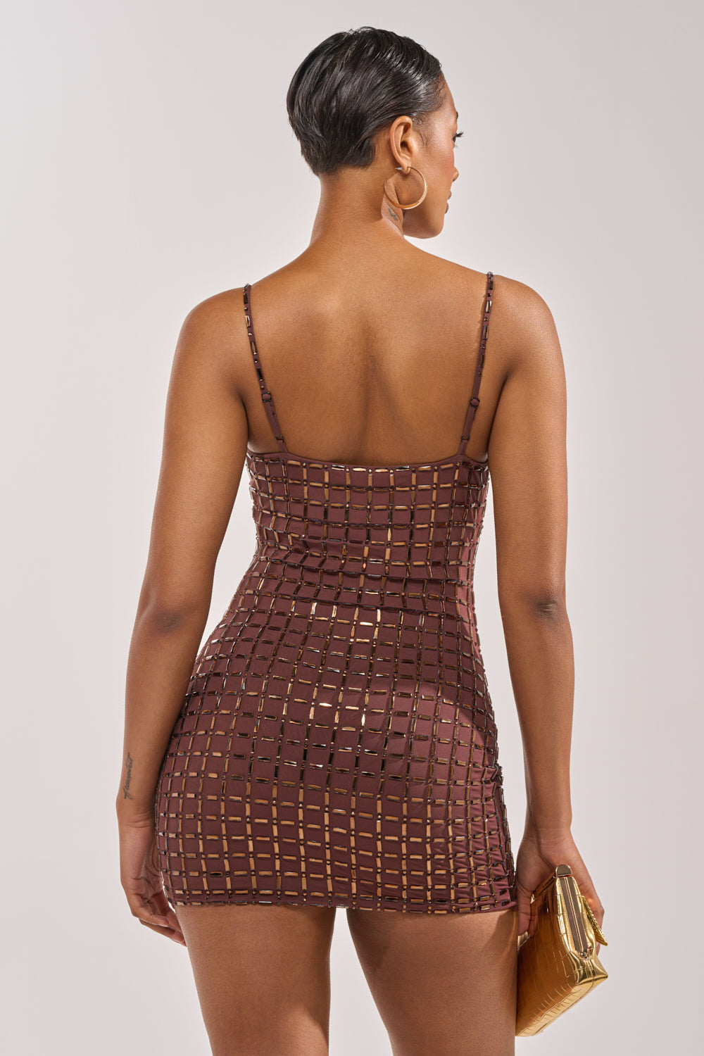 A woman with short hair and hoop earrings, wearing the DANCE THE NIGHT AWAY RHINESTONE MINI DRESS, with a square cutout pattern, stands facing away holding a small gold clutch. 4