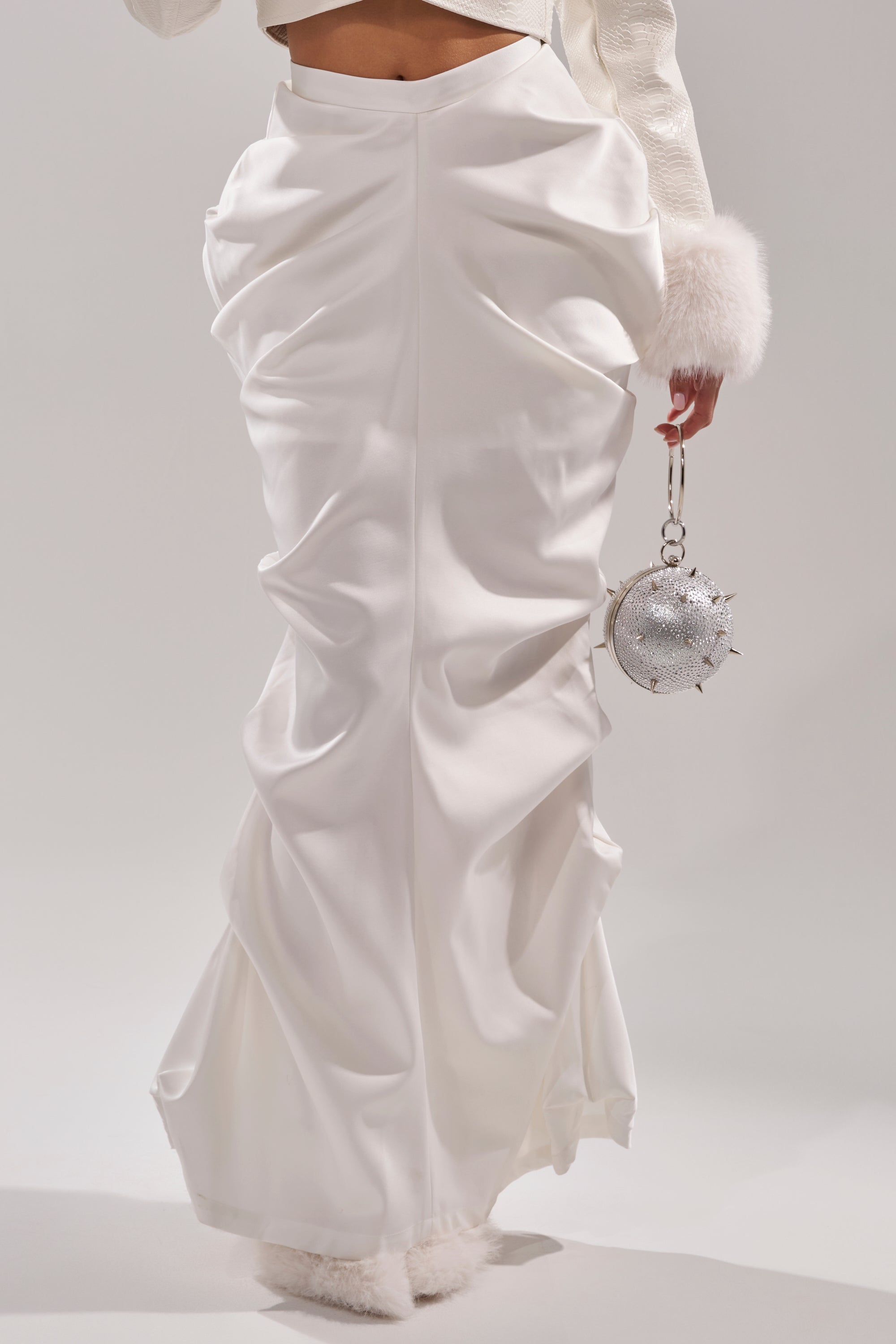 Wearing the FEELING THE VIBES SATIN MAXI SKIRT in white with ruched detailing, a cream jacket with fluffy cuffs, and carrying a small round silver spiked rhinestone handbag for a glamorous high-fashion look. 6