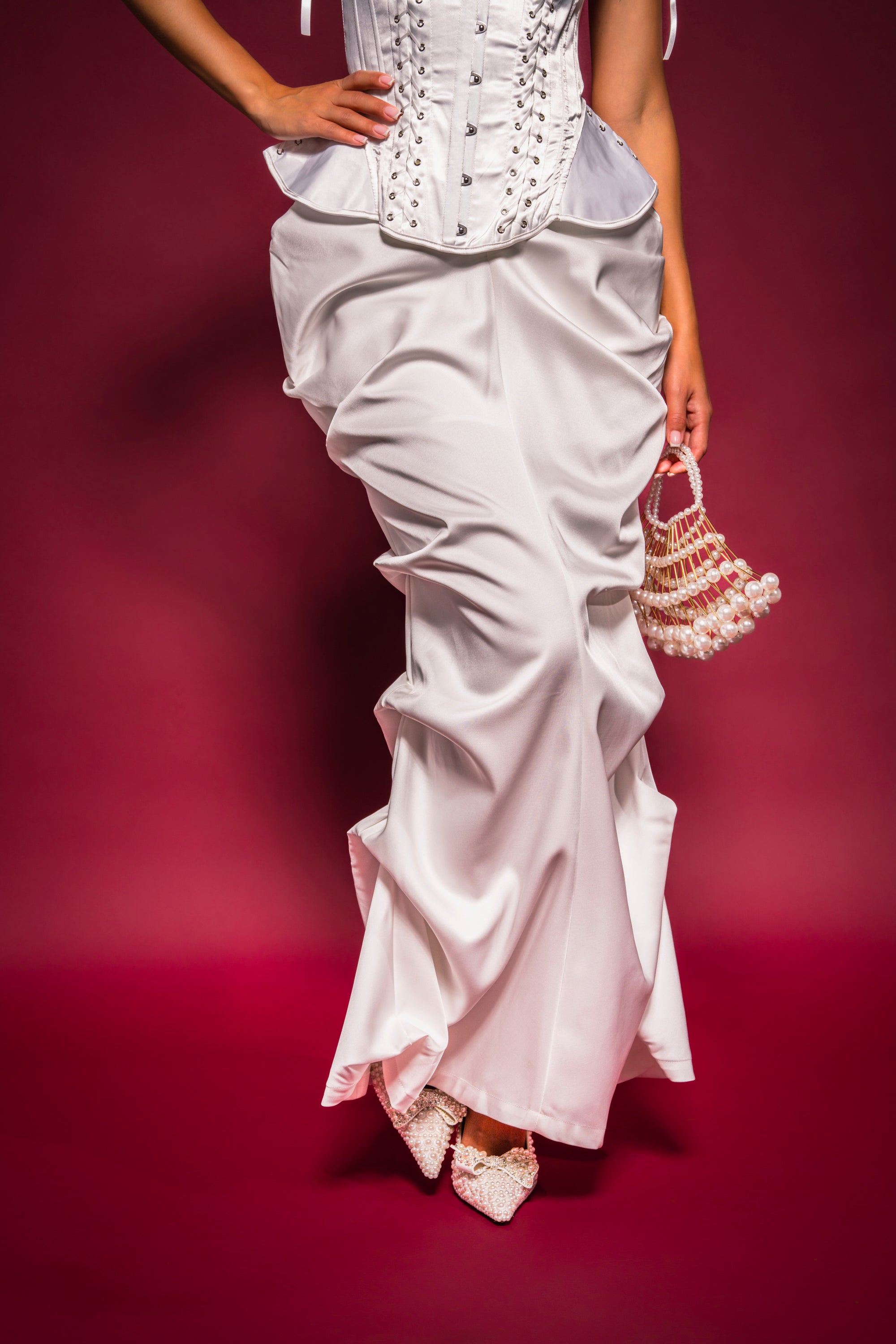 Wearing the FEELING THE VIBES SATIN MAXI SKIRT, a person in a white corset stands against a deep pink background, holding a beaded handbag and wearing pointed embellished shoes. 2
