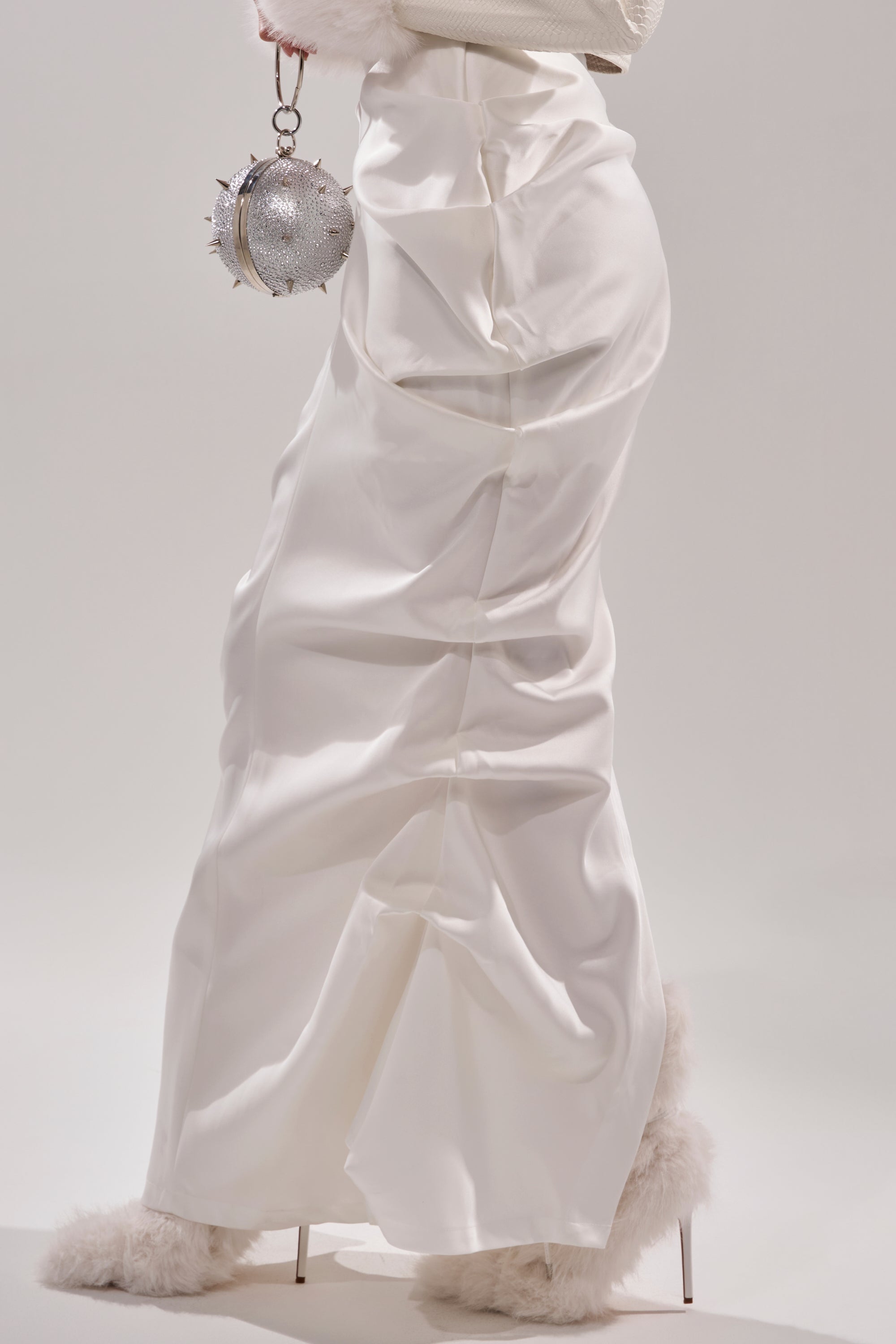 Someone wears the FEELING THE VIBES SATIN MAXI SKIRT in silky white with ruched details, fluffy white shoes, and carries a round silver spiked chain handbag against a plain light background. 8