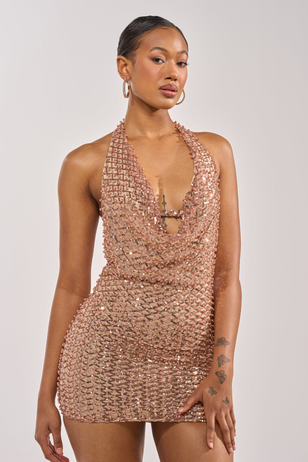 A woman poses confidently in the CHAMPAGNE RIOT RHINESTONE MINI DRESS, a sleeveless sequin mini with a cowl neckline. She accessorizes with hoop earrings, has slicked-back hair, and tattoos visible on her left hand and forearm. 0