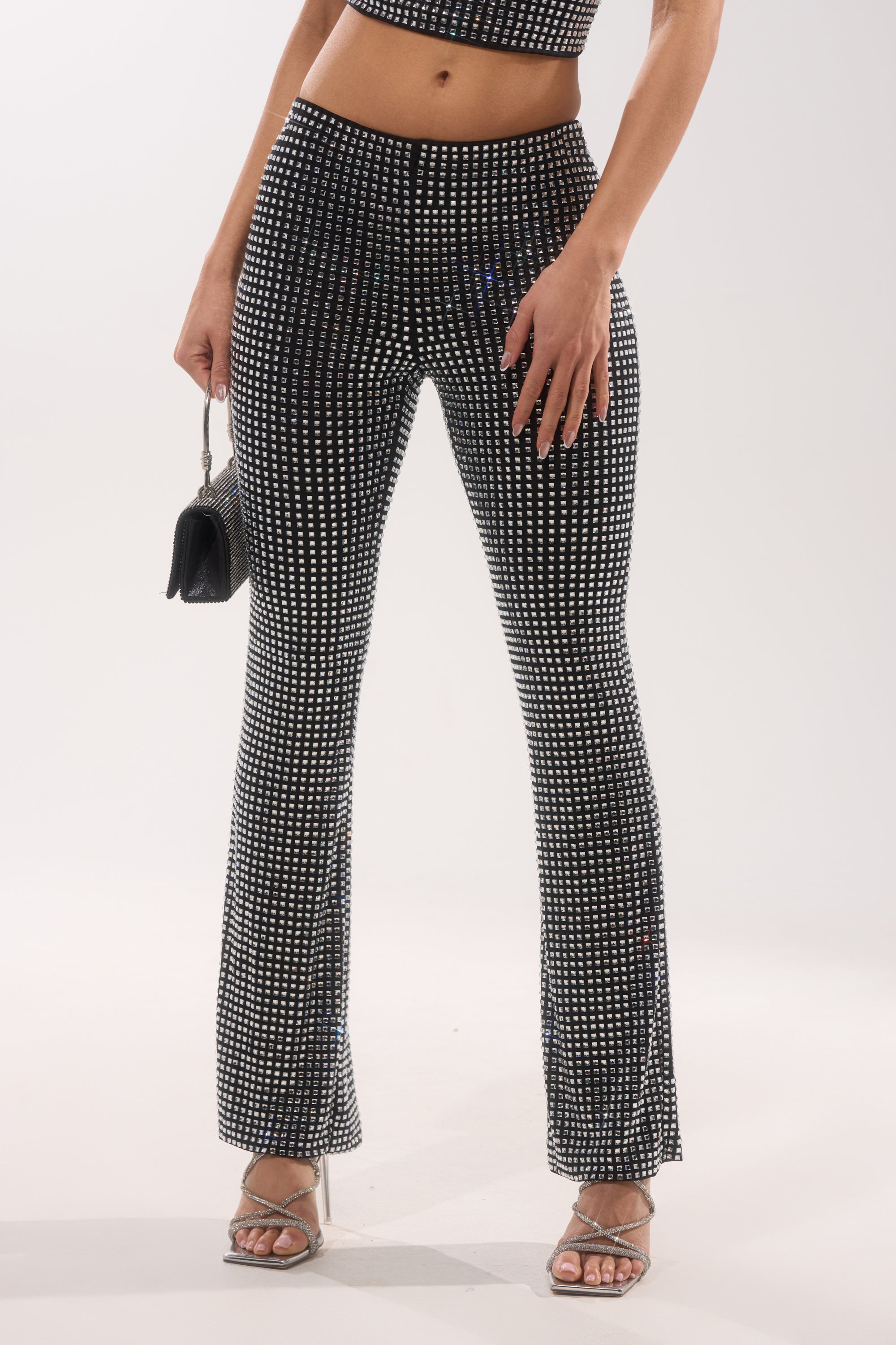 A woman poses against a white background in the ALL EYES ON ME RHINESTONE FLARE TROUSER with black and white rhinestone detail, a matching crop top, silver strappy heels, and a small black purse. 1