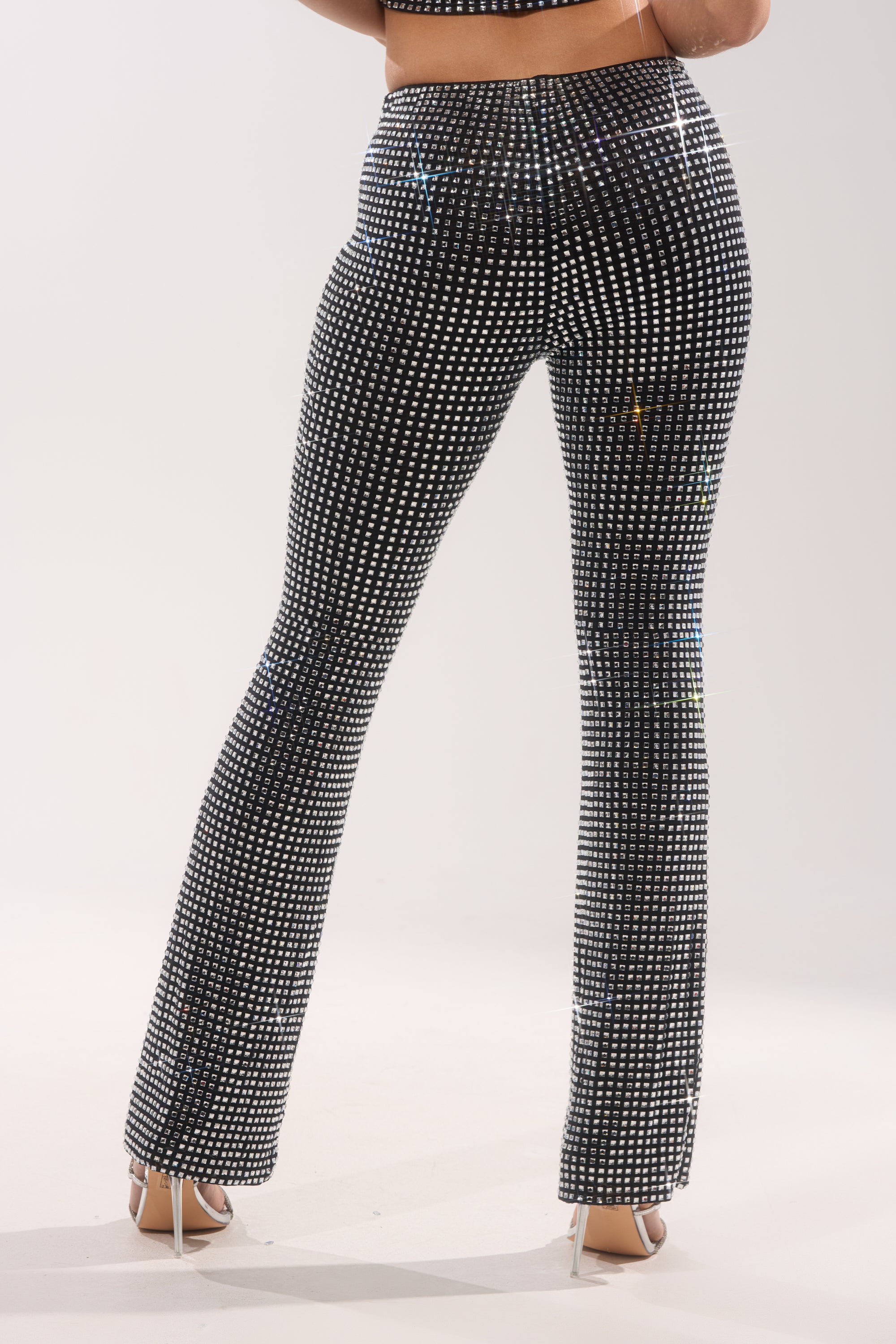 A person wearing ALL EYES ON ME RHINESTONE FLARE TROUSER—high-waisted black flares with a grid of reflective silver studs—paired with white high heels, seen from behind against a plain light background. 4
