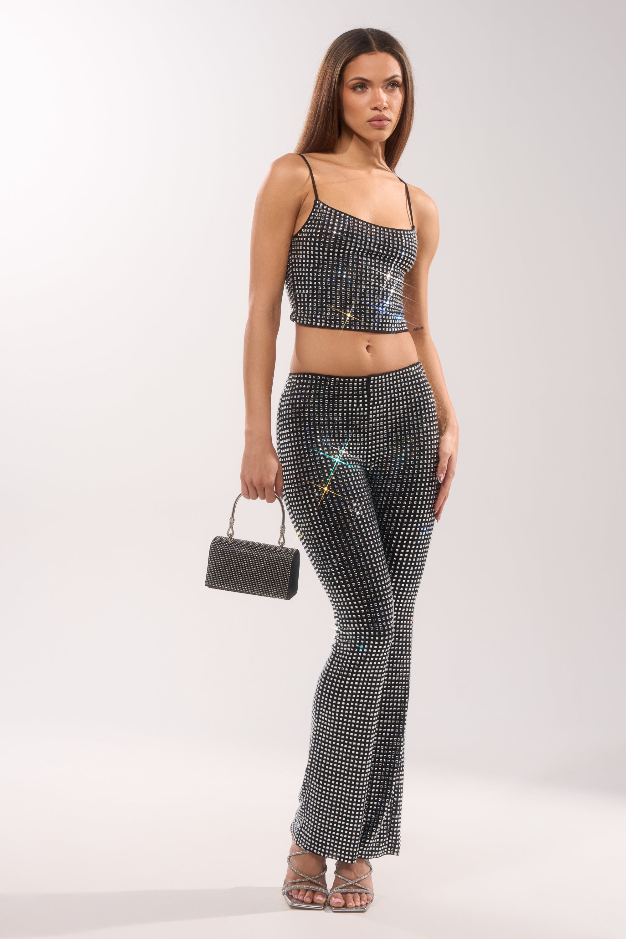 A woman stands against a plain background wearing sparkling ALL EYES ON ME RHINESTONE FLARE TROUSER, a rhinestone-studded black crop top, strappy heels, and holding a small glittery handbag. 5