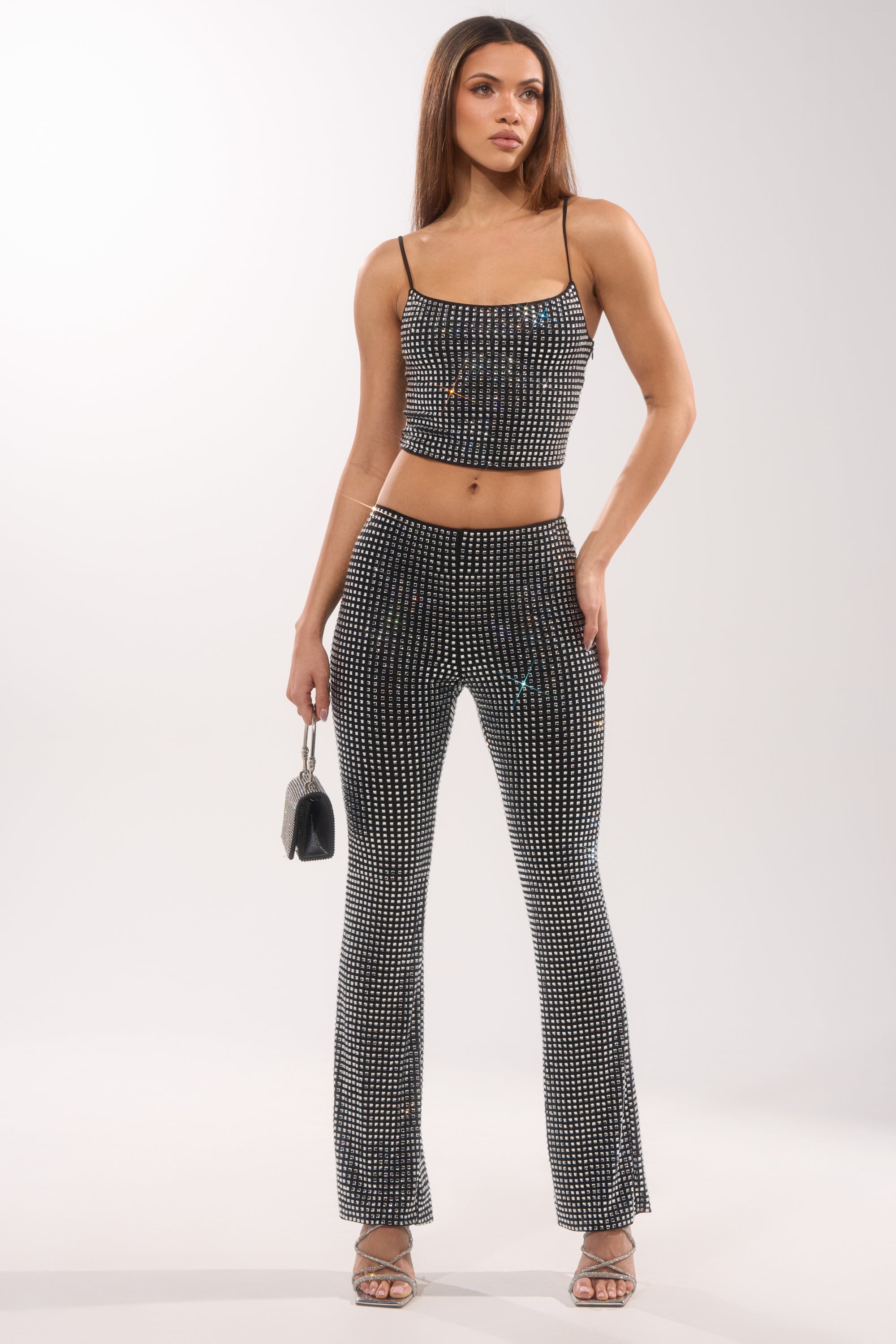 A woman poses in the ALL EYES ON ME RHINESTONE FLARE TROUSER, paired with a coordinating rhinestone crop top. She holds a small handbag, wears heeled sandals, and her straight, long brown hair stands out against a plain white background. 6