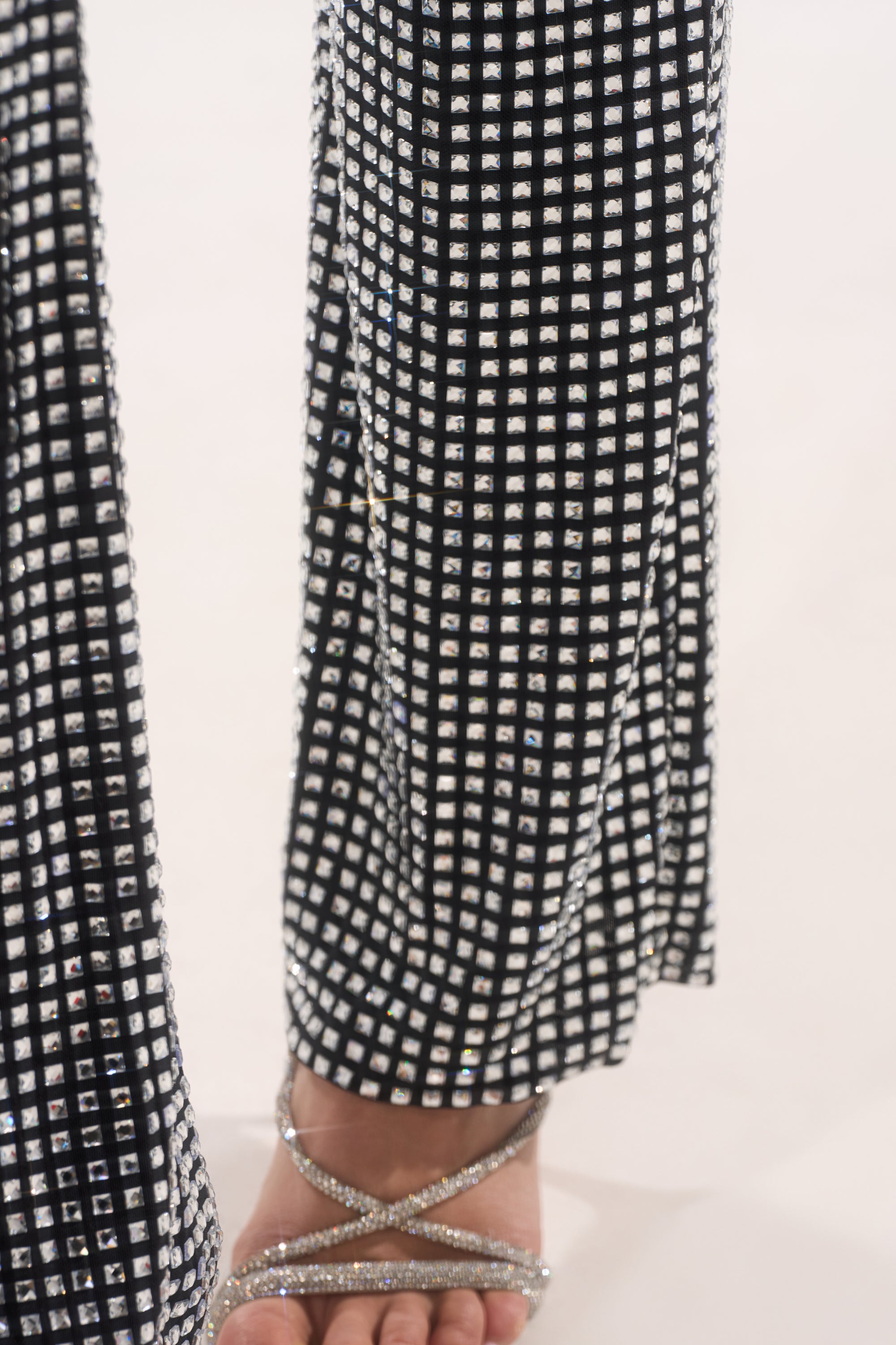 A close-up of a person’s lower legs wearing the ALL EYES ON ME RHINESTONE FLARE TROUSER, featuring a grid of sparkling rhinestones, paired with silver strappy heeled sandals on a light surface. 8